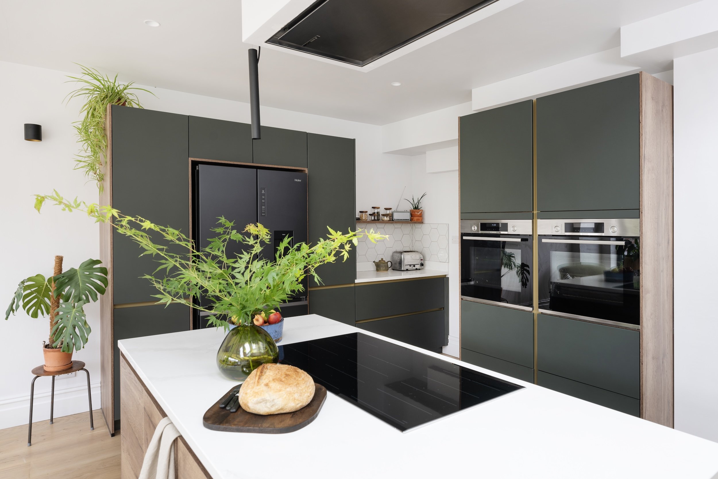 Handleless green kitchen with tall oak-effect units, integrated ovens and a white island with an induction hob, in North Bristol by JG Build.