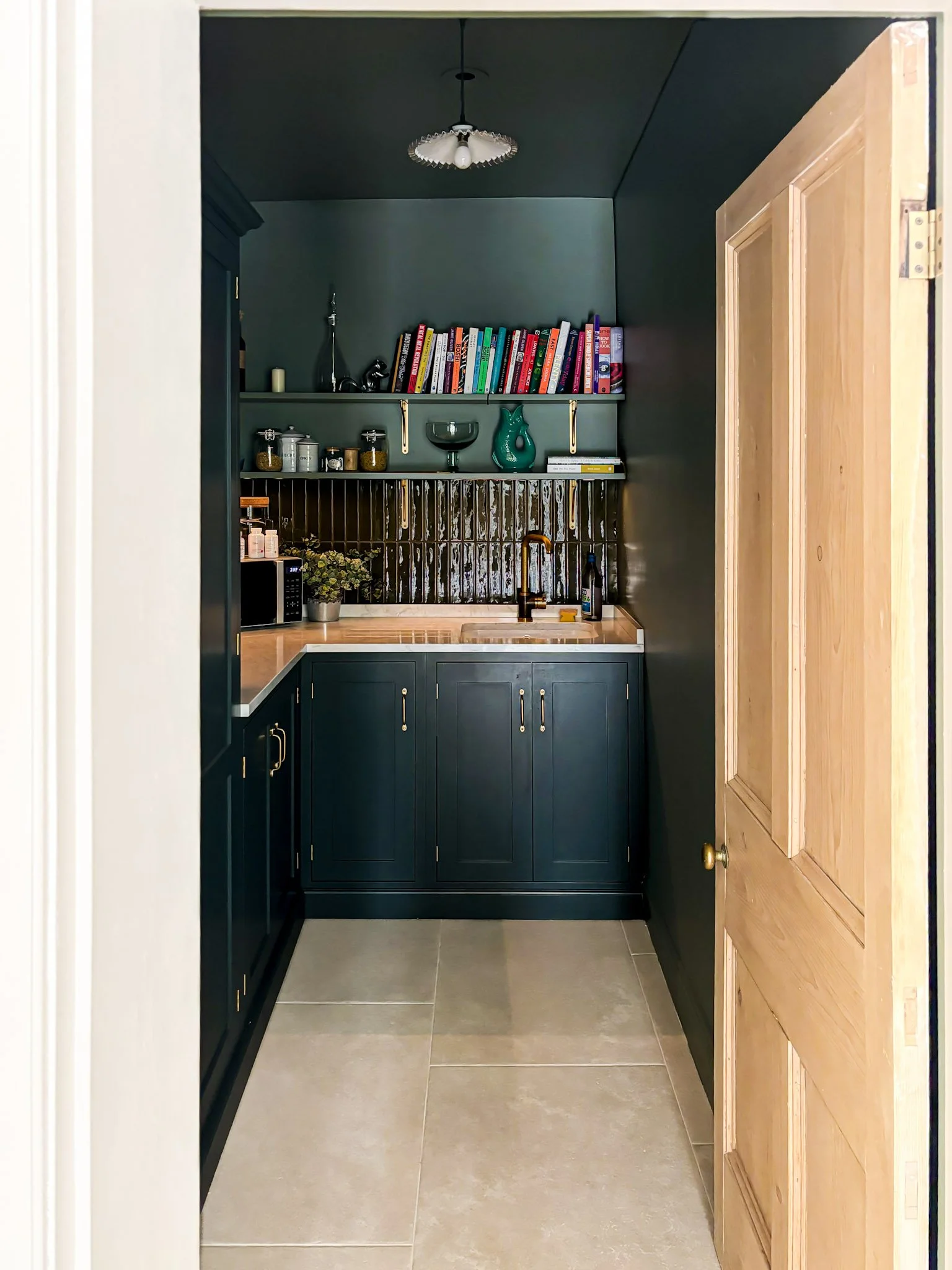 Larder with dark-painted cabinets and walls, a glazed tile splashback, open shelving and a small sink, seen through an original pine door, in Clifton by JG Build.