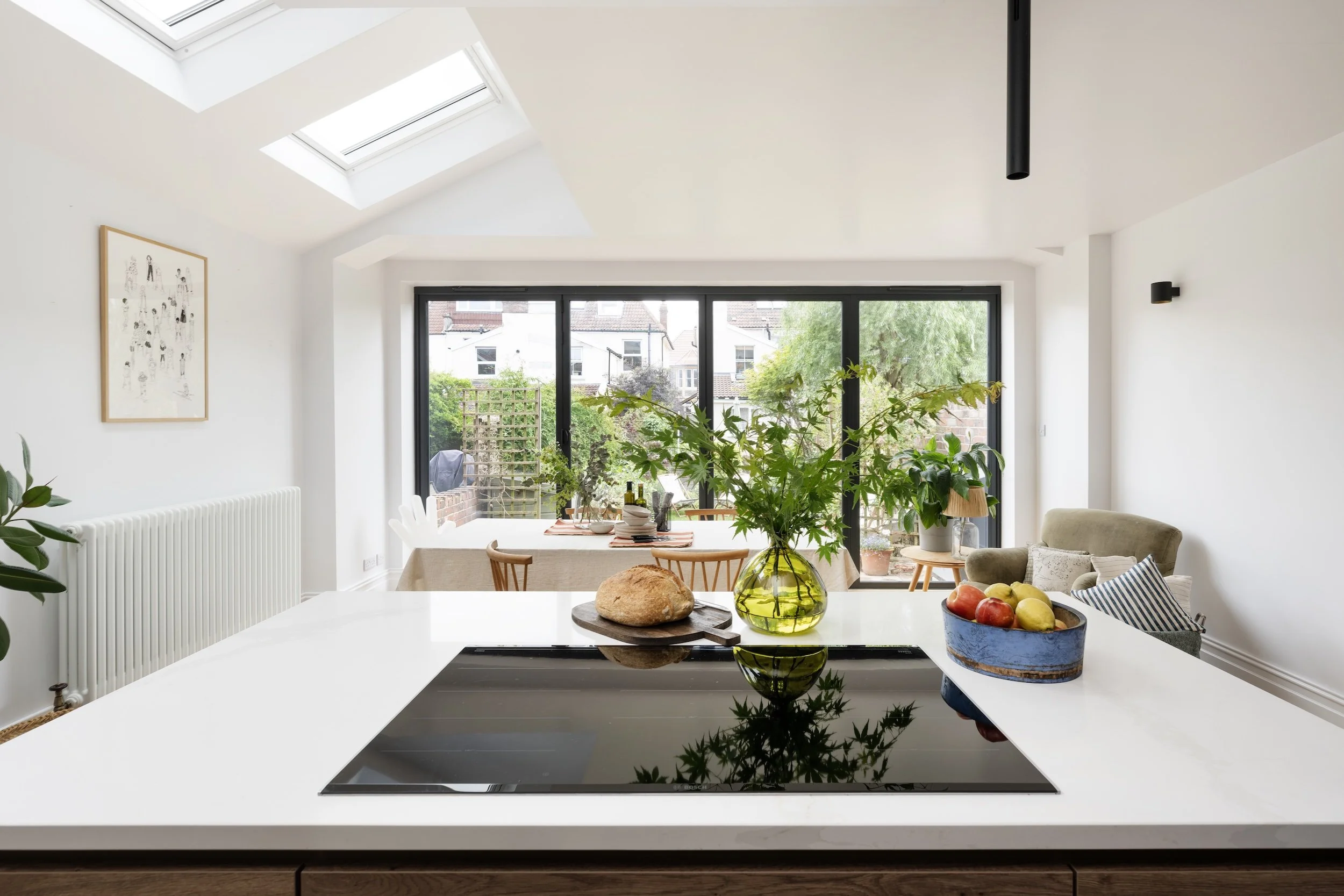 View across a white kitchen island with an induction hob towards the dining area, vaulted ceiling and bi-fold doors, in North Bristol by JG Build.