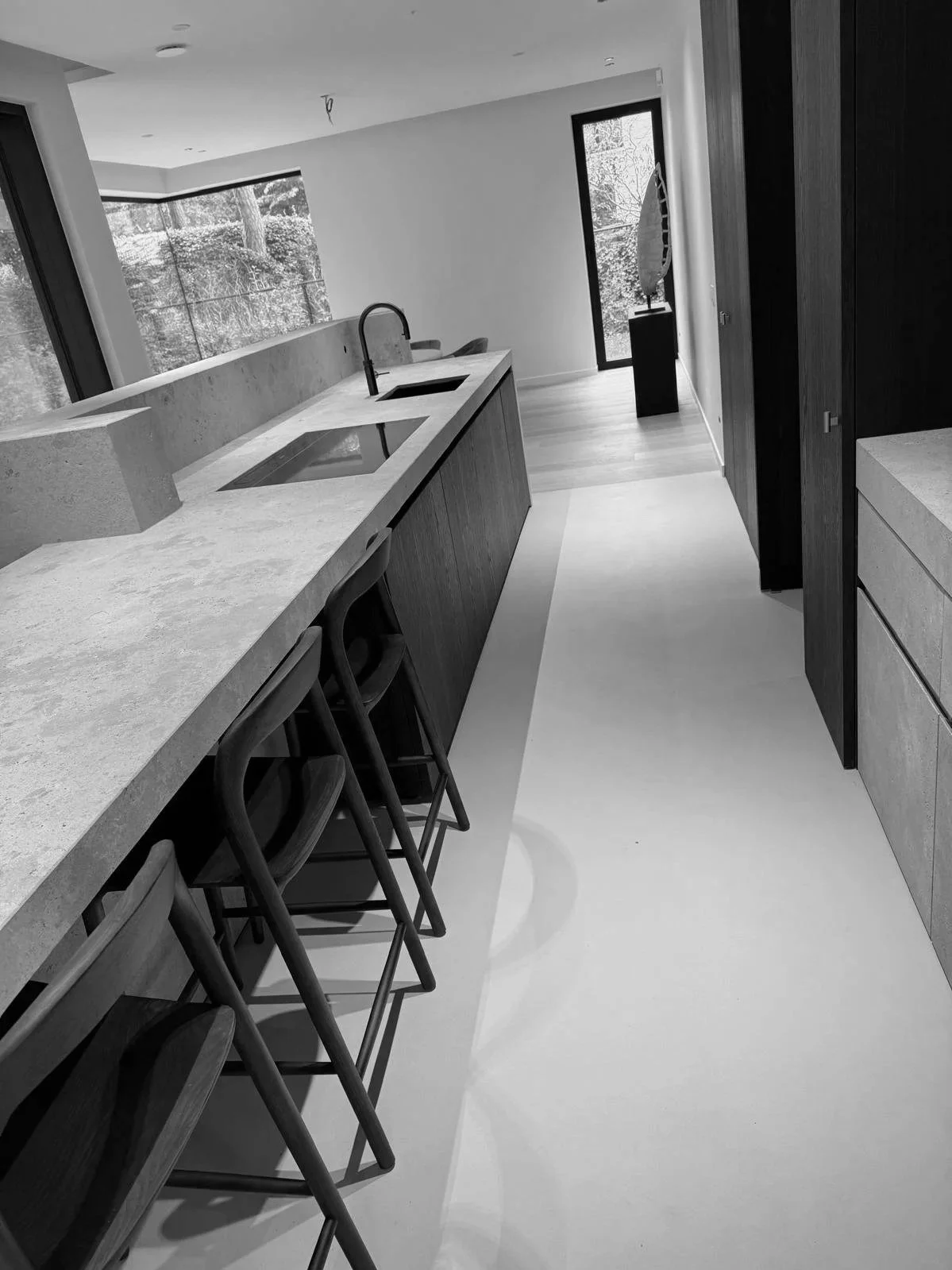Moderne keuken met een lange marmeren ontbijttafel, barkrukken, grote ramen met uitzicht op bomen, en een minimalistisch interieur.