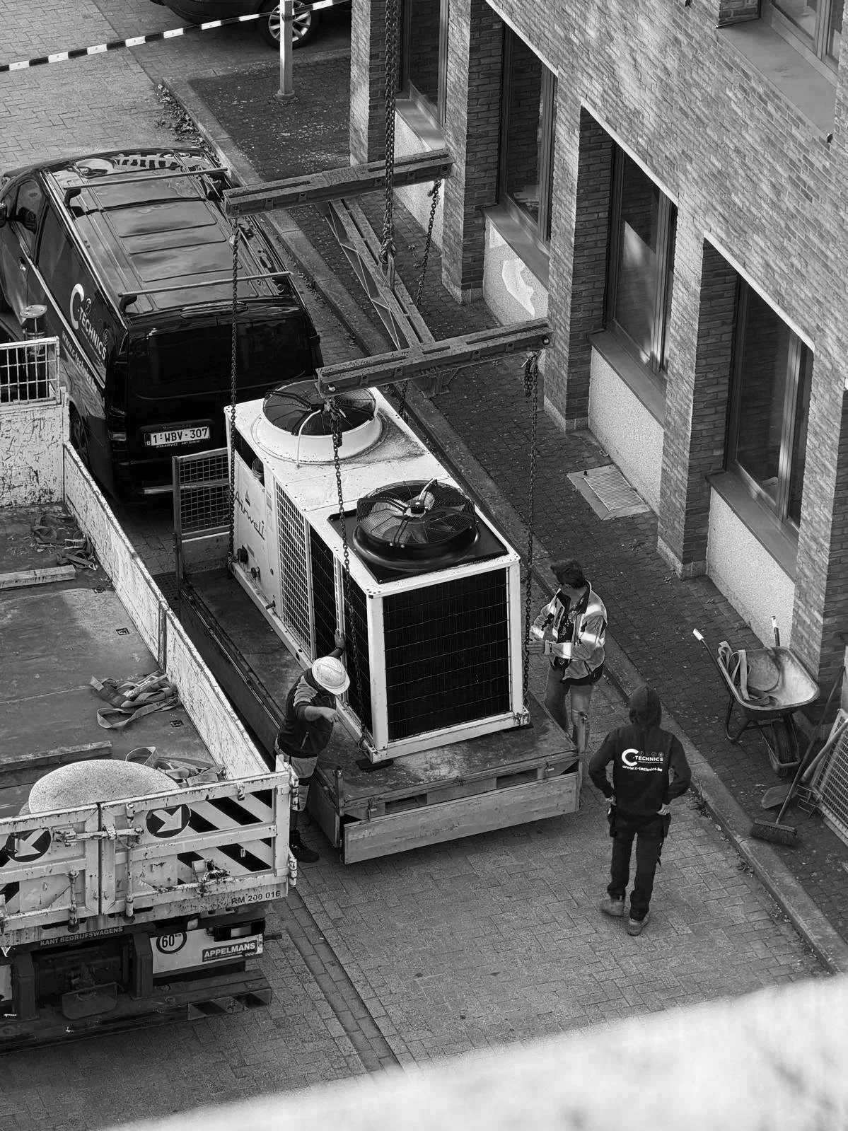 Een zwart-witte foto van een bouwplaats met twee werknemers die werken aan de installatie van een groot koelunit op een dak van een gebouw. Een personenwagen en een vrachtwagen staan in de buurt.