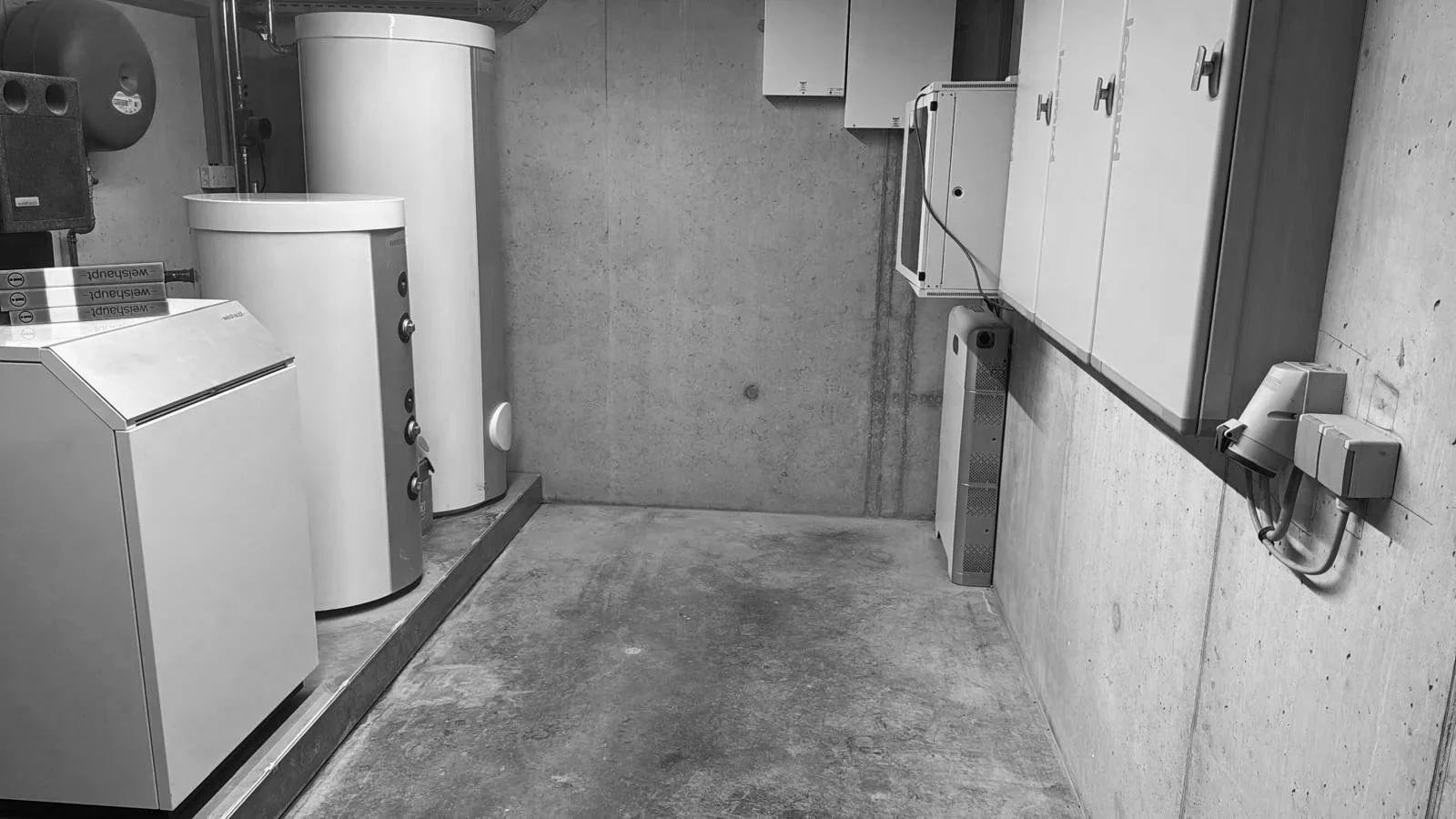 Een zolderruimte met waterketels, een wasdroogcombinatie en verwarmingsradiatoren, met betonwanden en vloer.