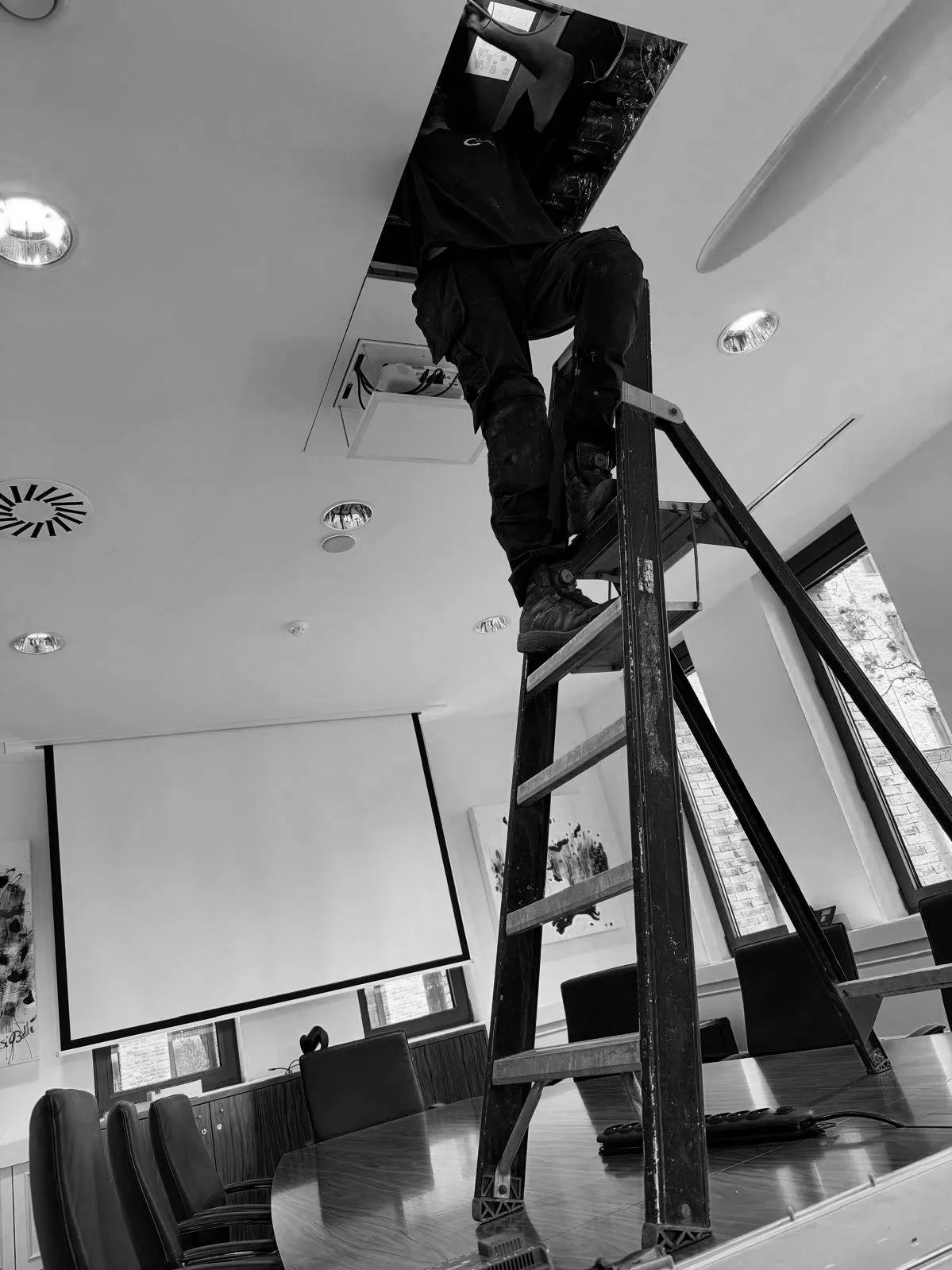 Een persoon staat op een ladder en werkt op het plafond van een vergaderruimte, mogelijk met technische of onderhoudswerkzaamheden.