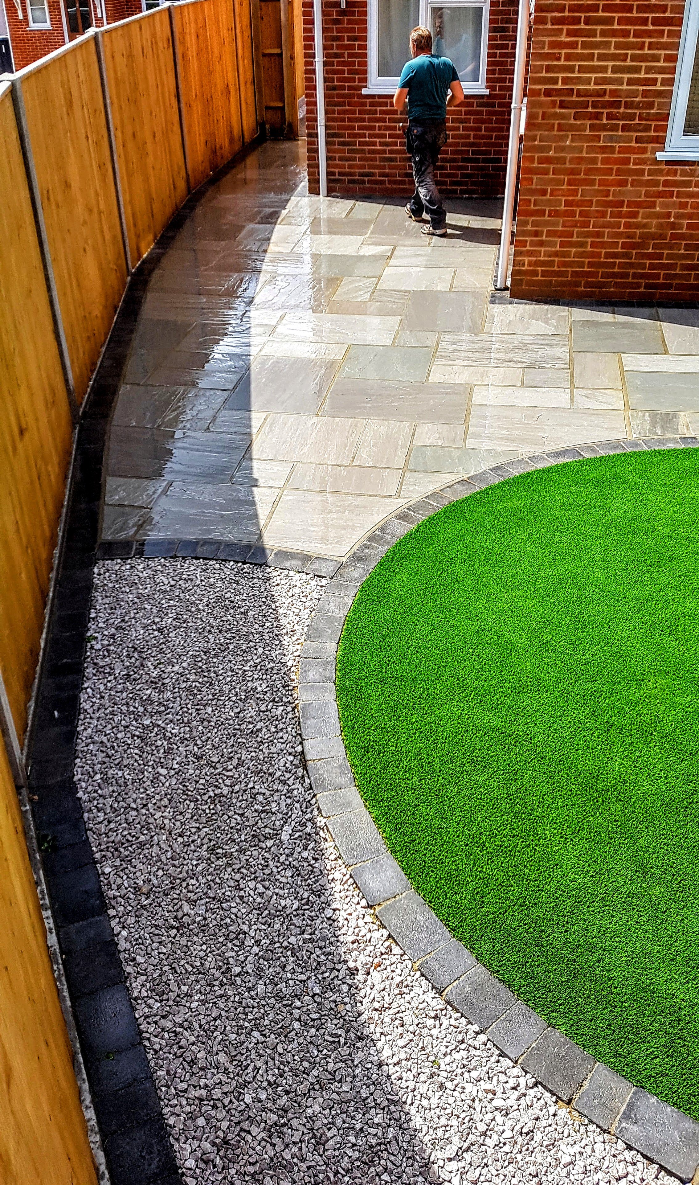 A backyard with a curved pathway made of large stone tiles, a section of gravel, and a bright green artificial grass lawn. A wooden fence lines the left side, and a brick house with a window is in the background. A person is walking along the house, 