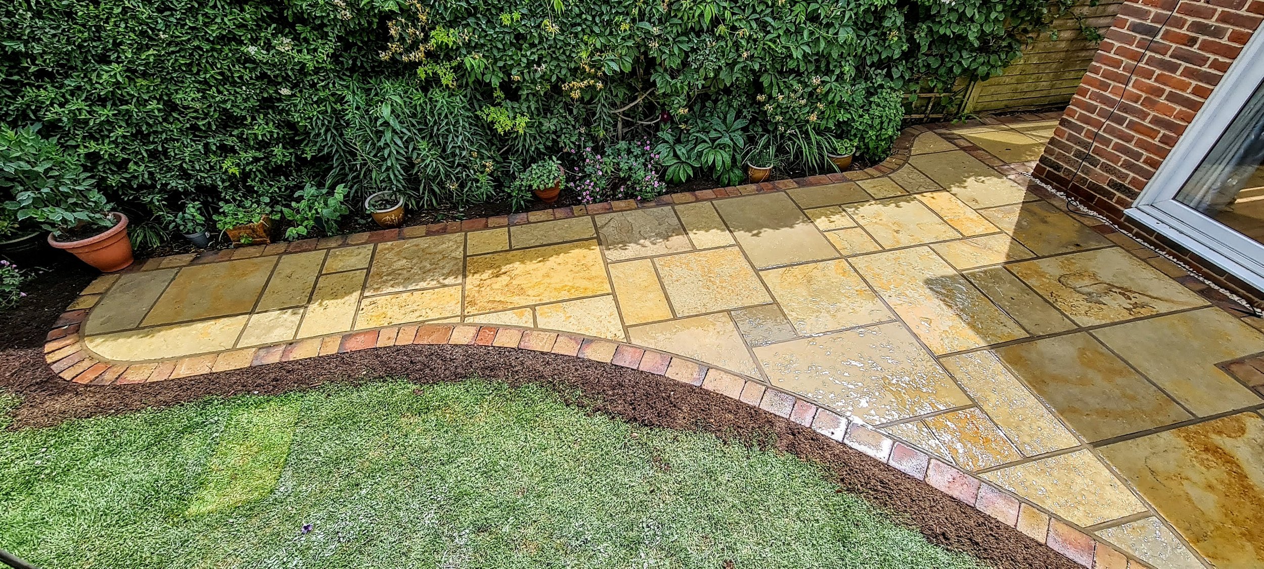 Wet yellow and tan stone patio with brick border beside a lawn, along with a background of green foliage and potted plants.