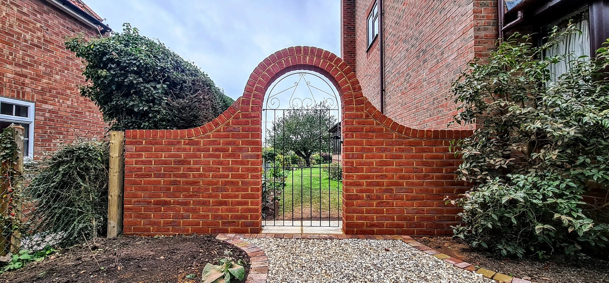 A brick wall with a decorative arch and gate leading to a backyard garden with green grass, trees, and shrubs.