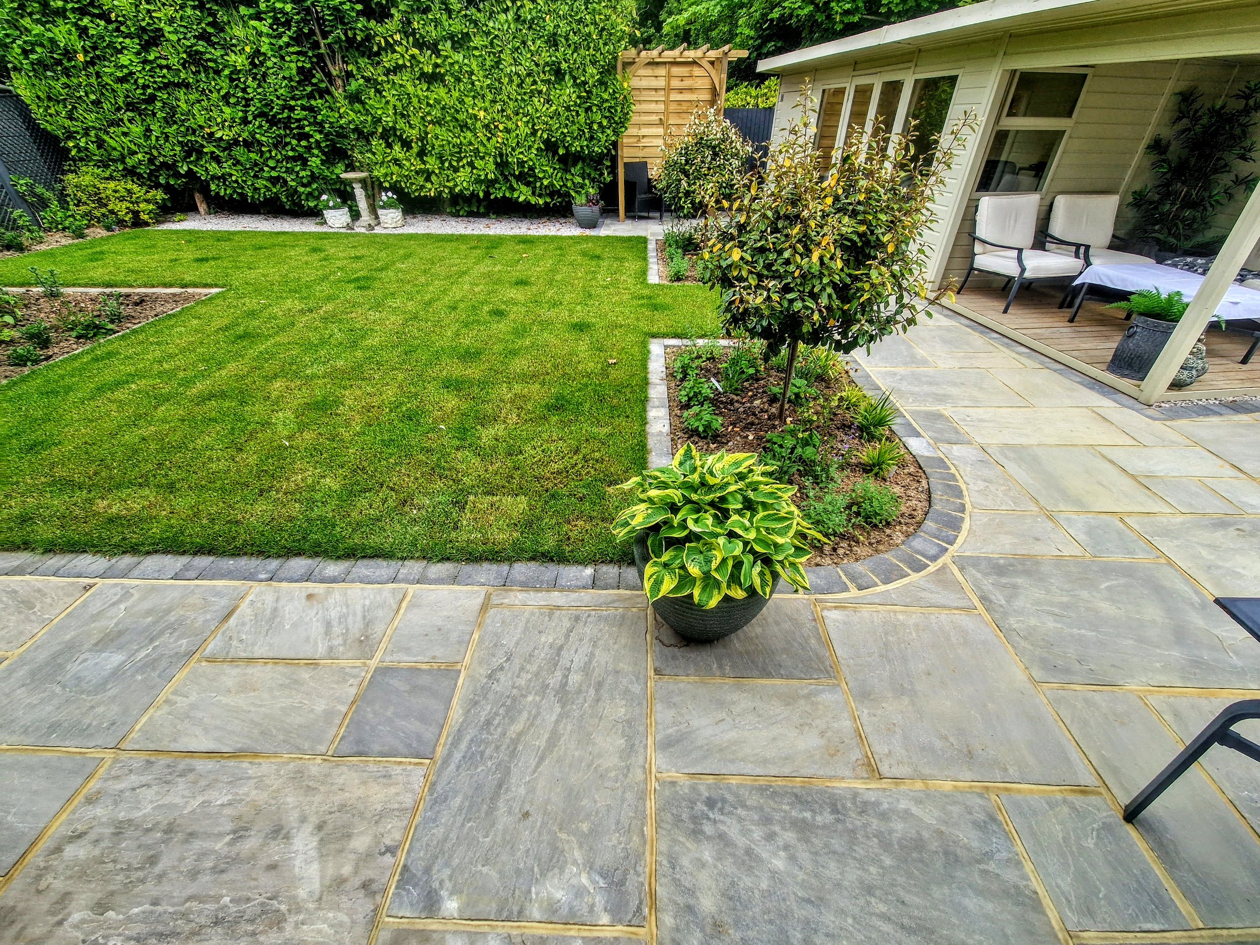 A backyard with a green lawn, potted plants, a stone patio, a white house with a porch, outdoor chairs, and lush green fencing.