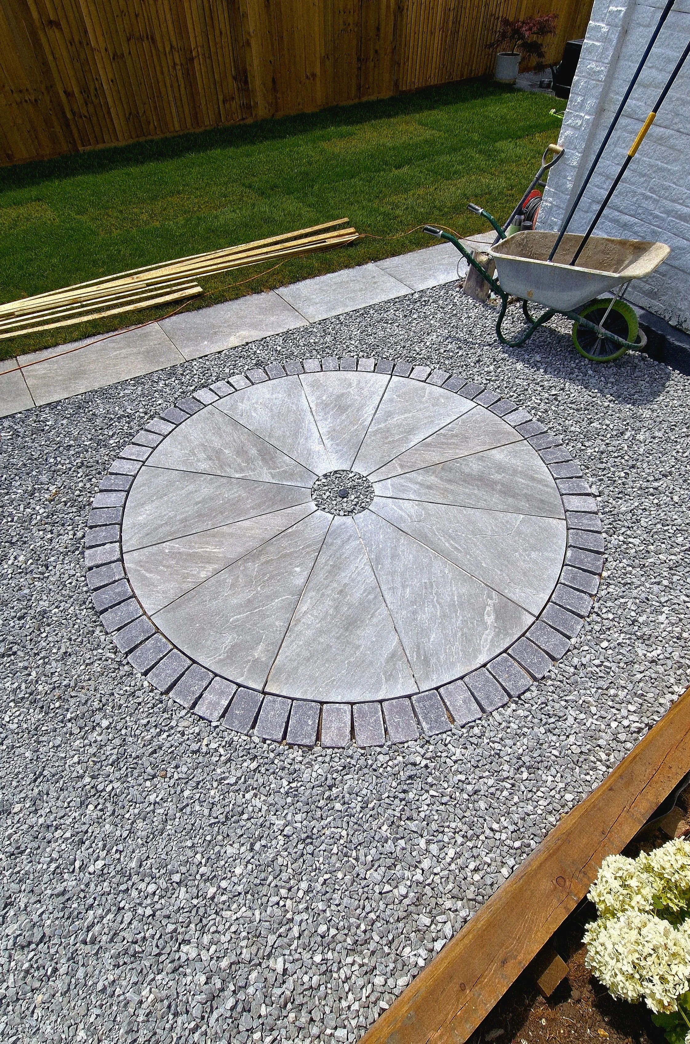 Garden patio with a circular stone pattern, gravel surrounding, a wheelbarrow, garden tools, and a wooden fence and greenery in the background.