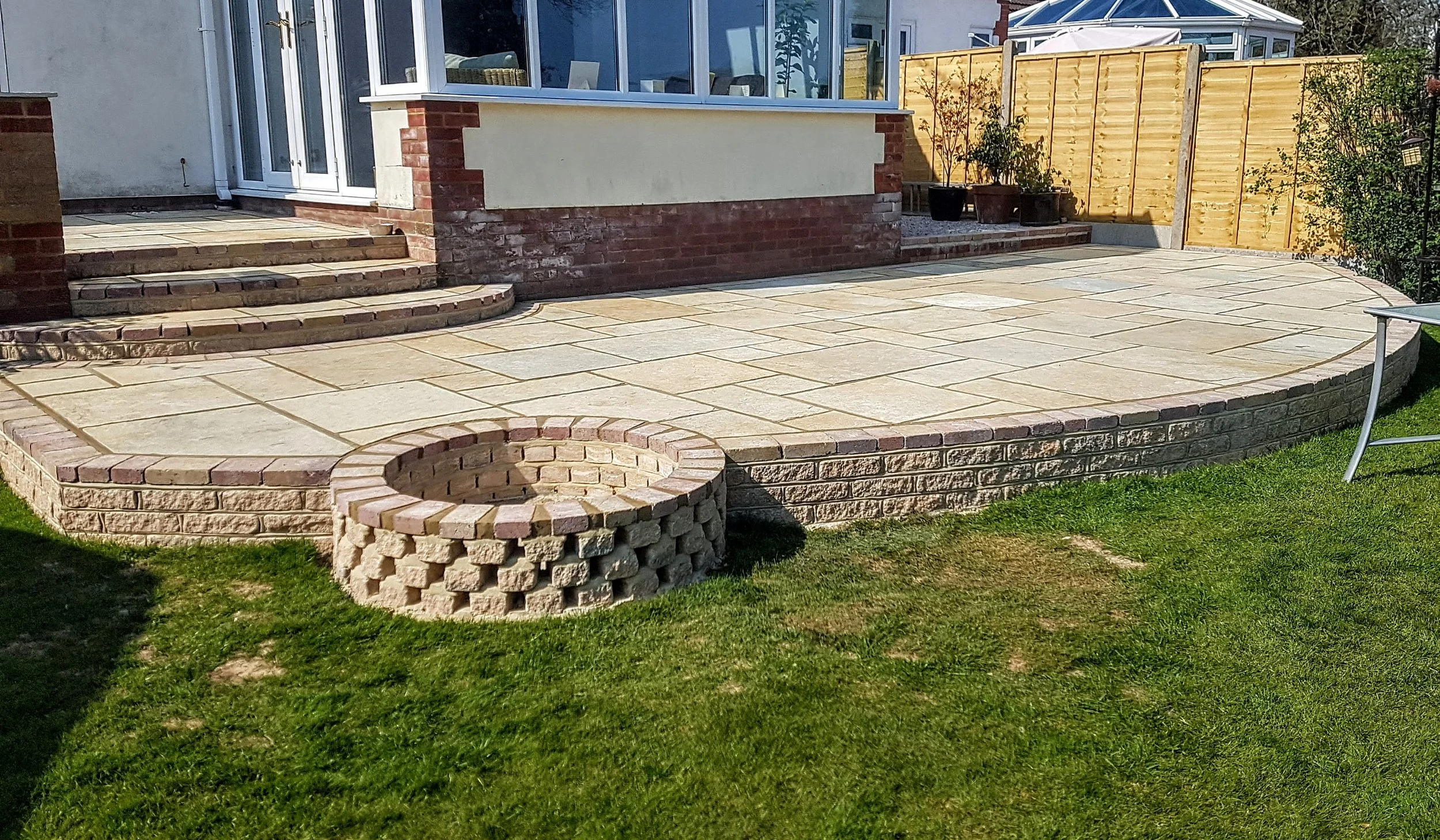 Backyard patio with a curved staircase leading to a sliding glass door, a fire pit made of bricks, and a fenced garden area with potted plants.