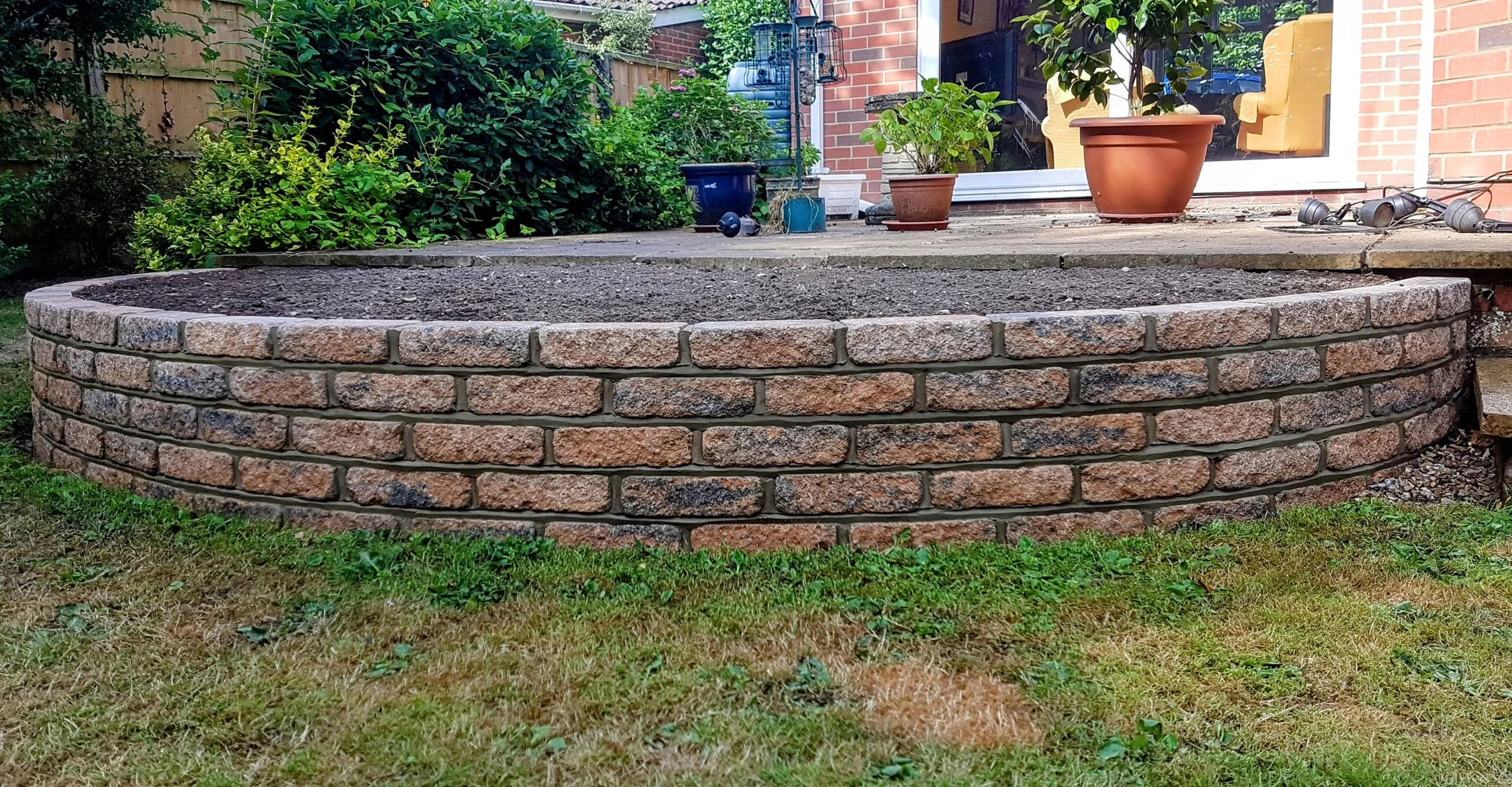 A raised garden bed made of brick with soil inside, surrounded by green grass and plants, with potted plants and gardening tools nearby.