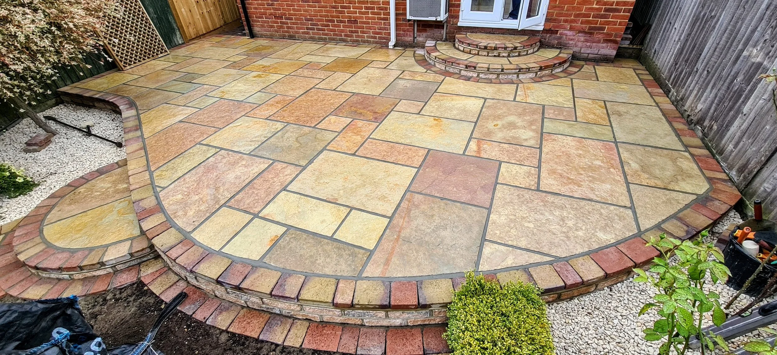 A newly constructed patio made of multi-colored stone tiles with curved brick edges surrounding a small set of brick steps leading to a door. The patio is enclosed by wooden fences on two sides.