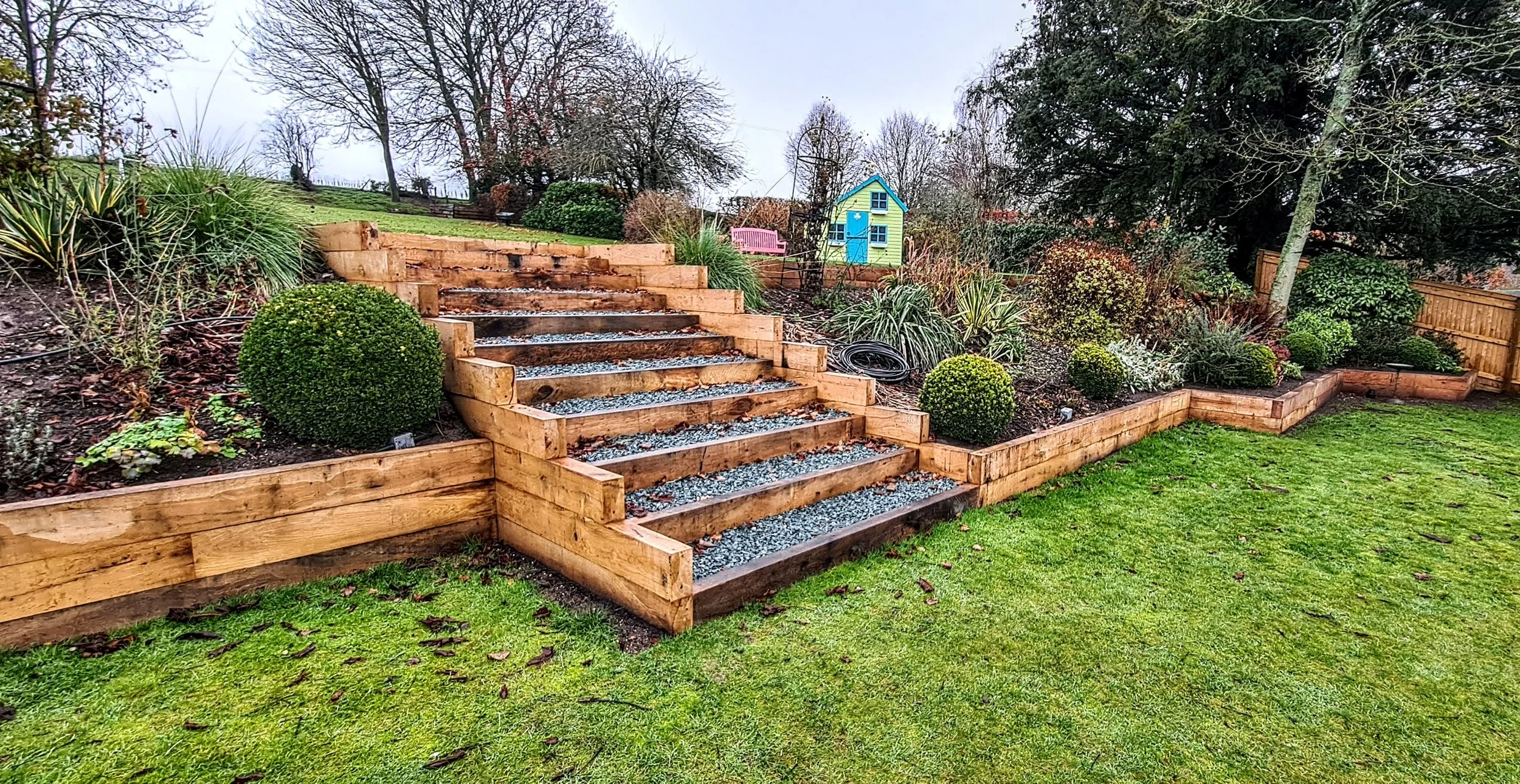 A landscaped backyard garden with wooden steps leading up a slope, surrounded by various green bushes and plants. In the background, there are trees, a pink bench, a small yellow playhouse with a blue door, and a wooden fence.