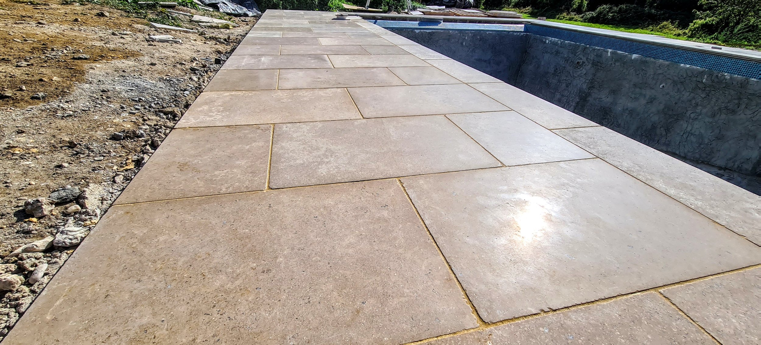 A partially completed outdoor patio with large beige pavers, next to a swimming pool with dark interior, and some construction debris and dirt on the side.