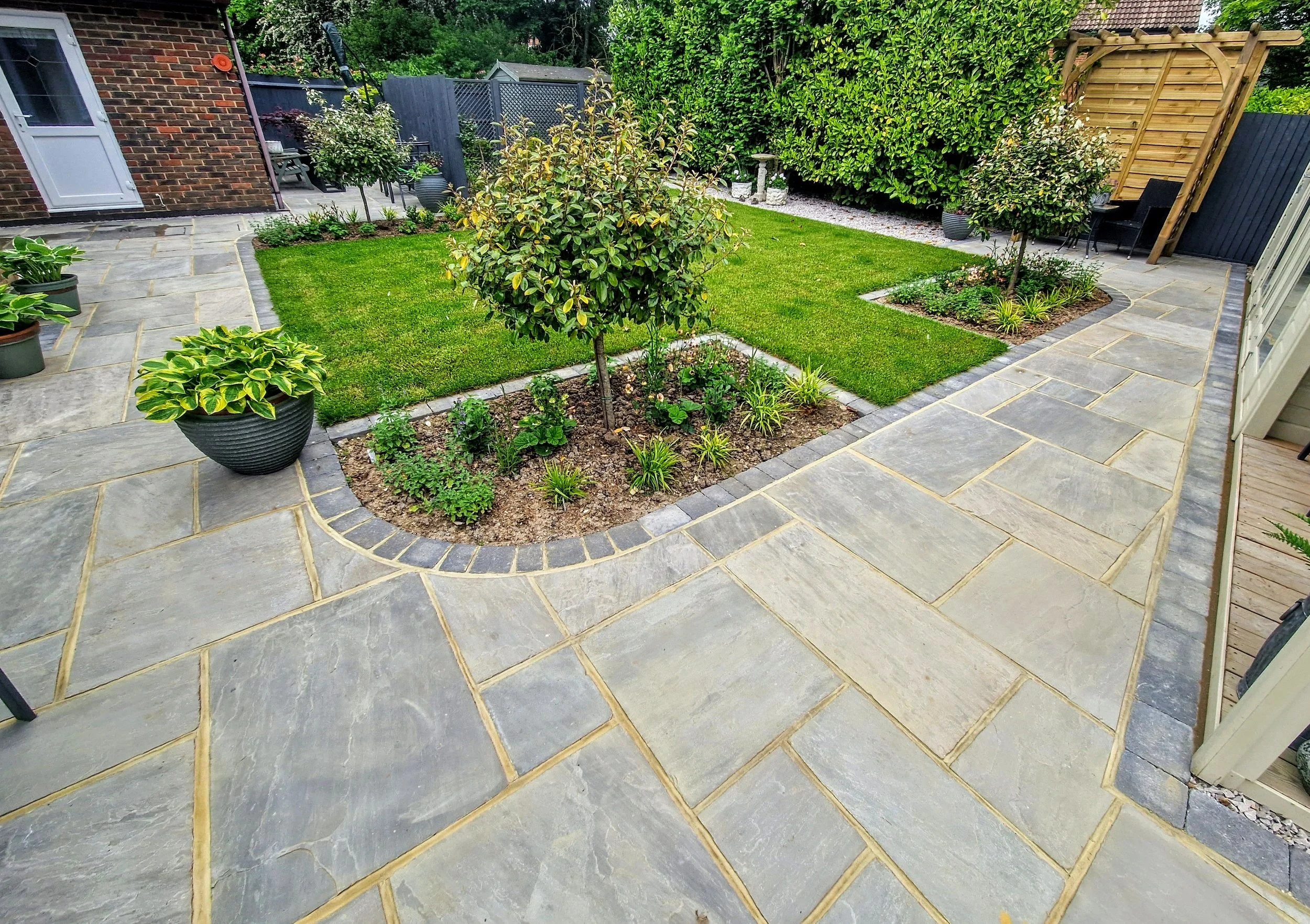 A backyard garden with a stone patio, green grass, small trees, potted plants, and a privacy fence.