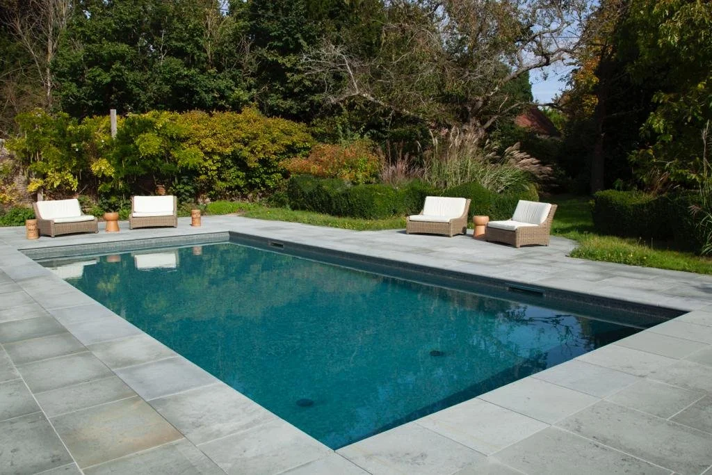A backyard with a rectangular swimming pool, surrounded by gray stone paving, and outdoor seating with white cushions and small terracotta tables. There are trees and shrubs in the background.