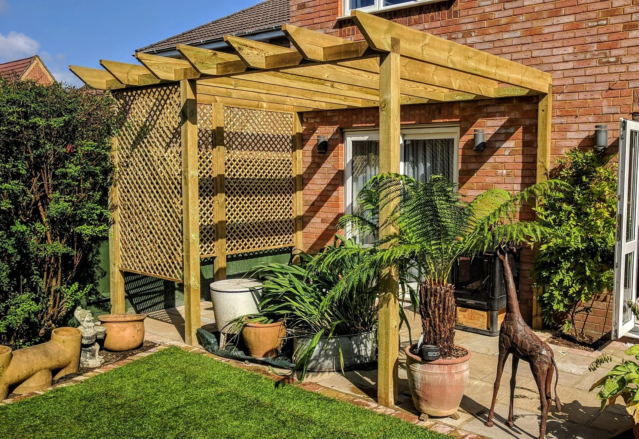 A backyard patio with an unfinished wooden pergola, potted plants, and garden decorations next to a brick house with a sliding door and outdoor lights.