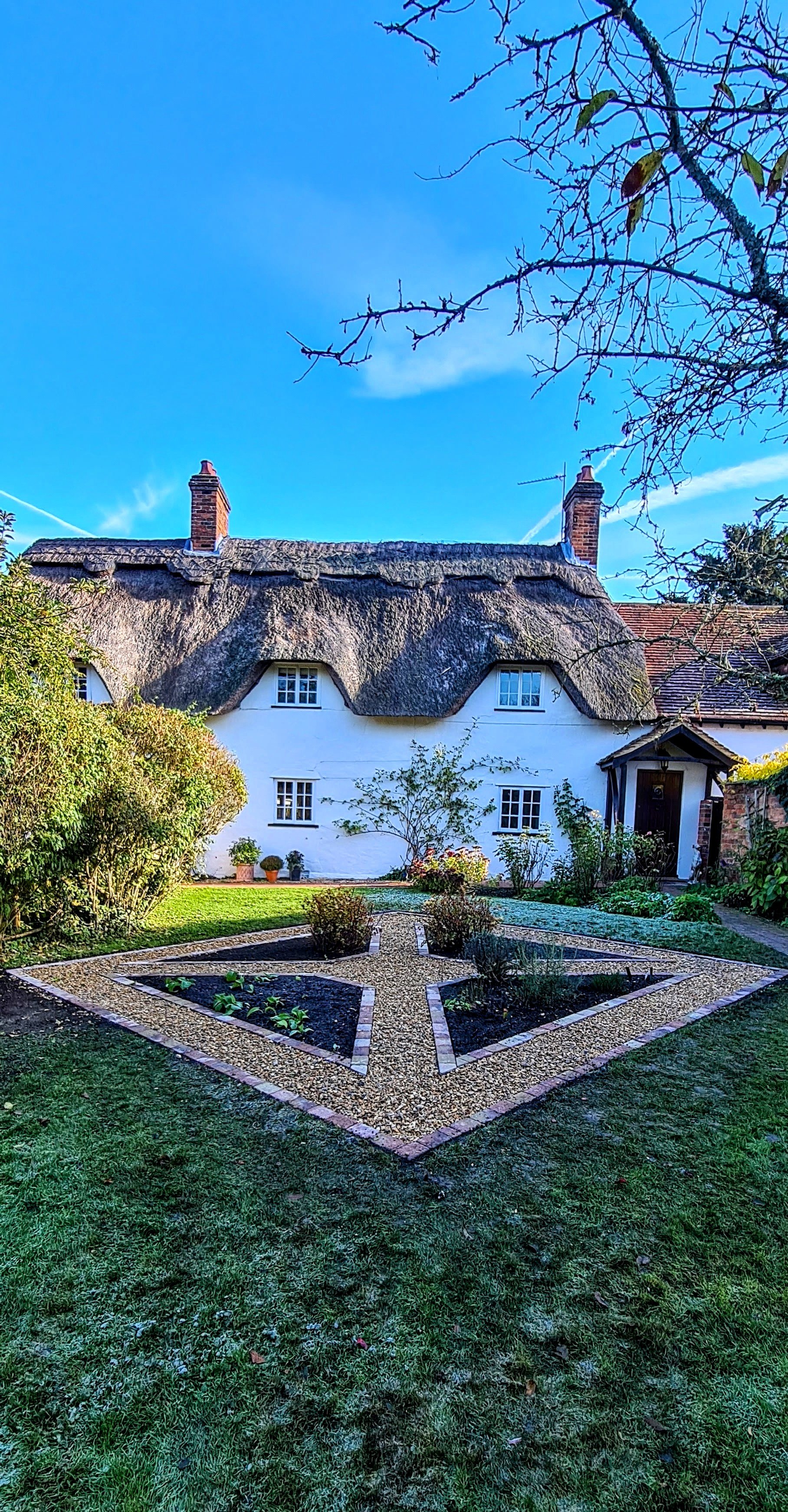 A quaint white cottage with a thatched roof, surrounded by a garden with neatly planted flower beds, bushes, and trees, under a clear blue sky.