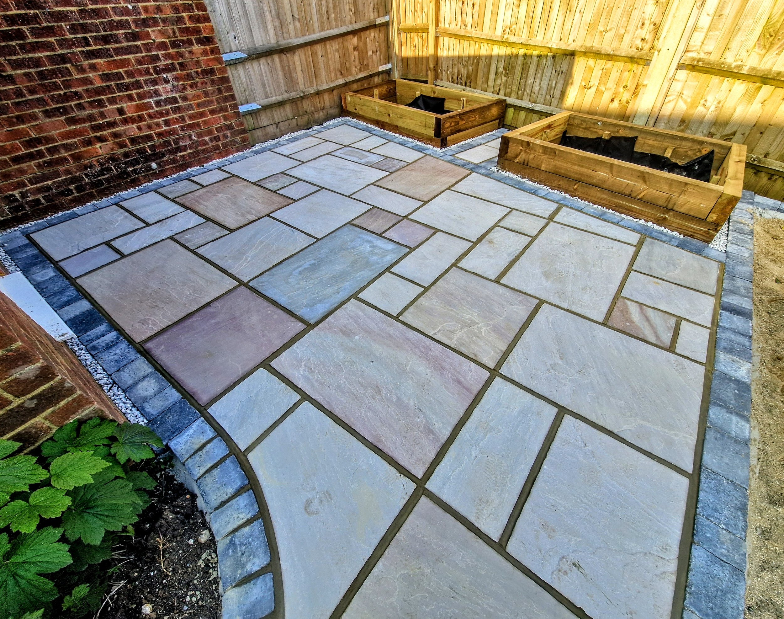 A small outdoor patio area with gray stone pavers, two wooden garden beds with black liner, a brick wall on the left, and a wooden fence on the right and back.