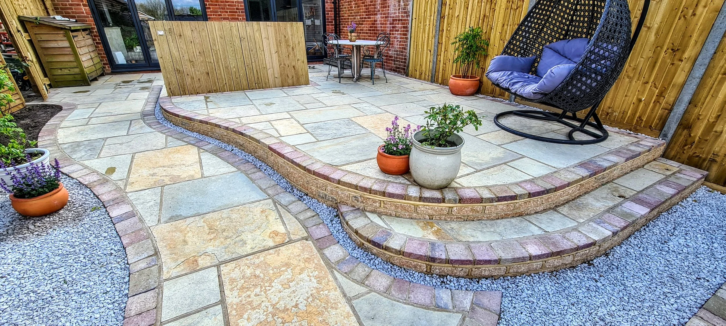 Private backyard patio with flagstone flooring, brick and wooden fencing, potted plants, and outdoor seating including a hanging egg chair with cushions and a table with chairs.
