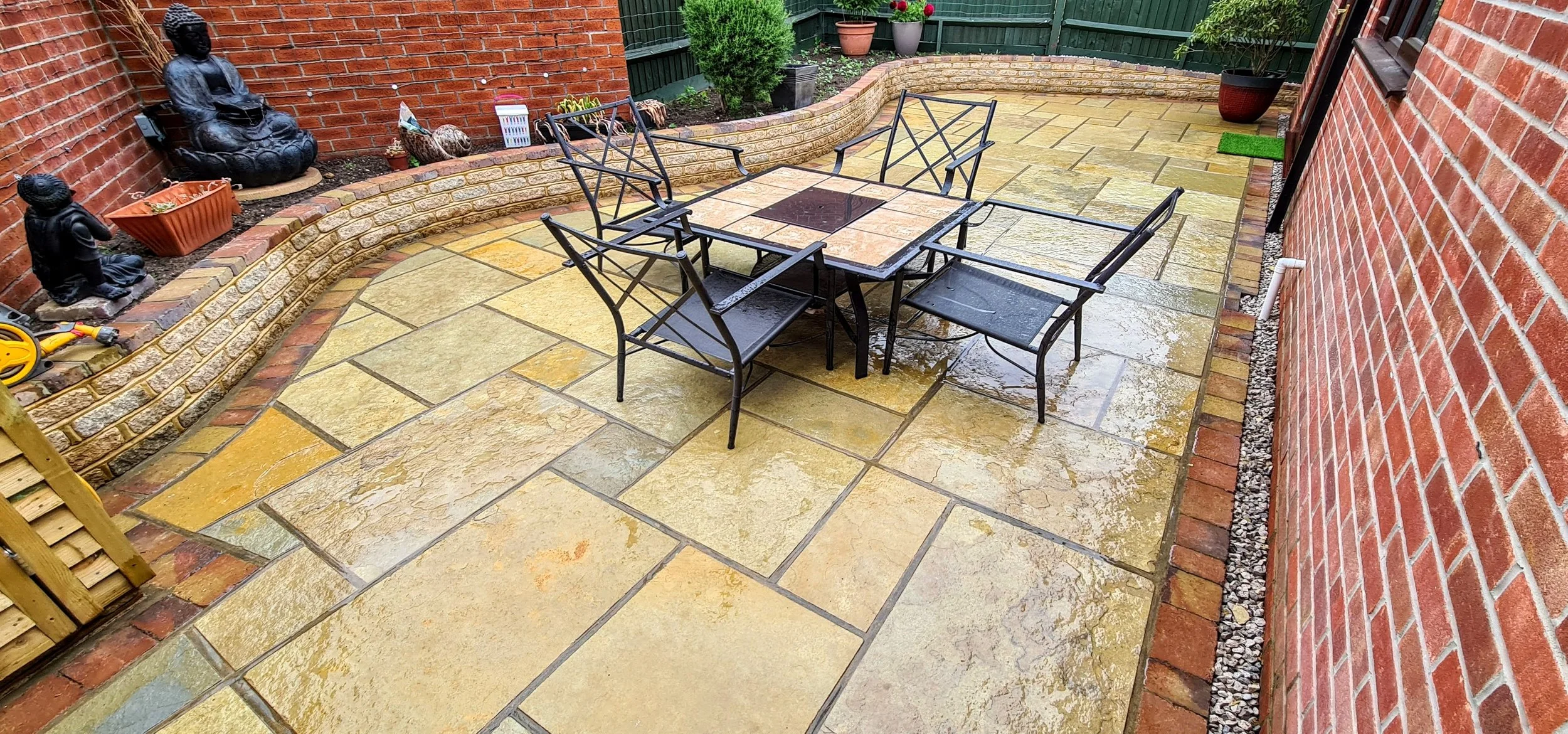 A wet outdoor patio with a dining table and six black chairs, surrounded by brick walls, potted plants, garden statues, and a rain-soaked tiled floor.