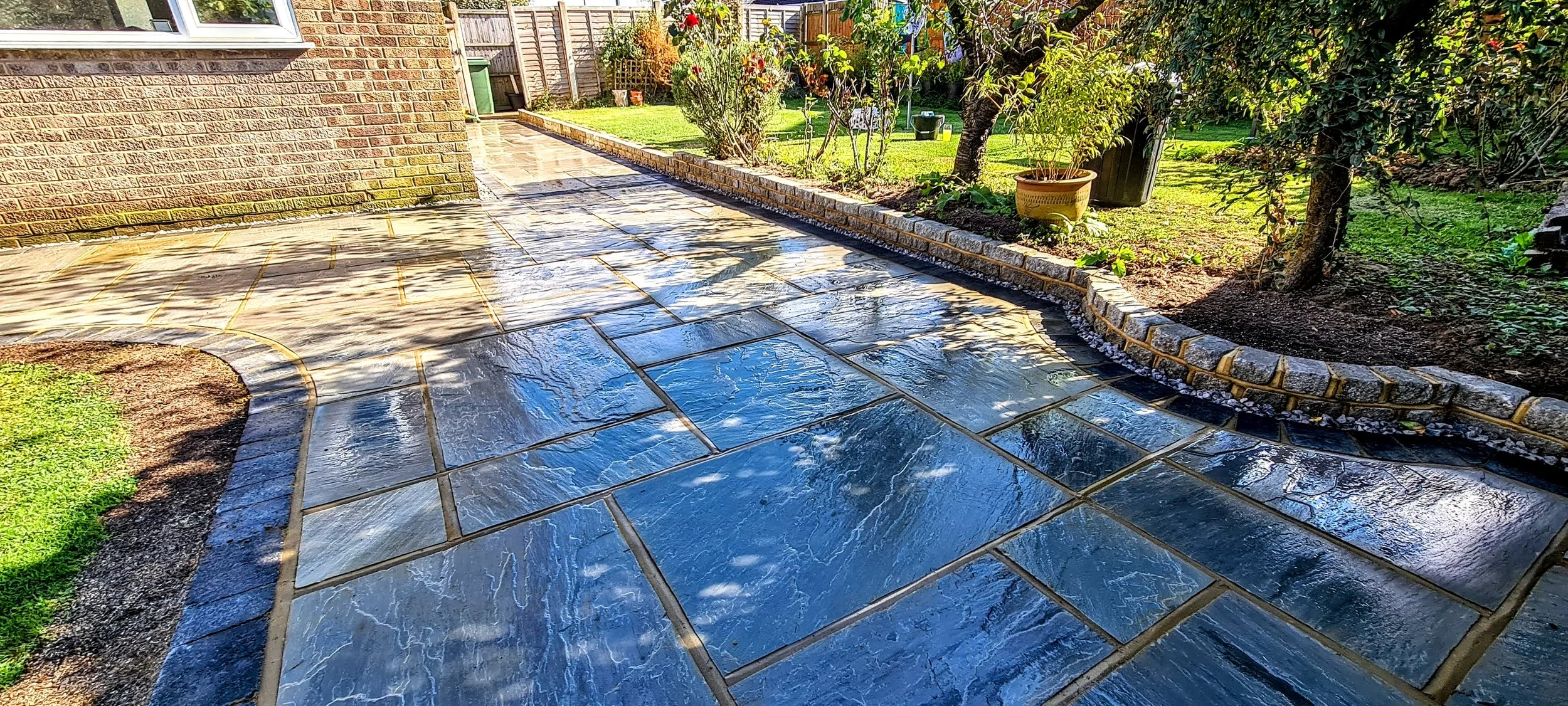 Freshly sealed stone patio in backyard with greenery, trees, and flowerbed along the edge.