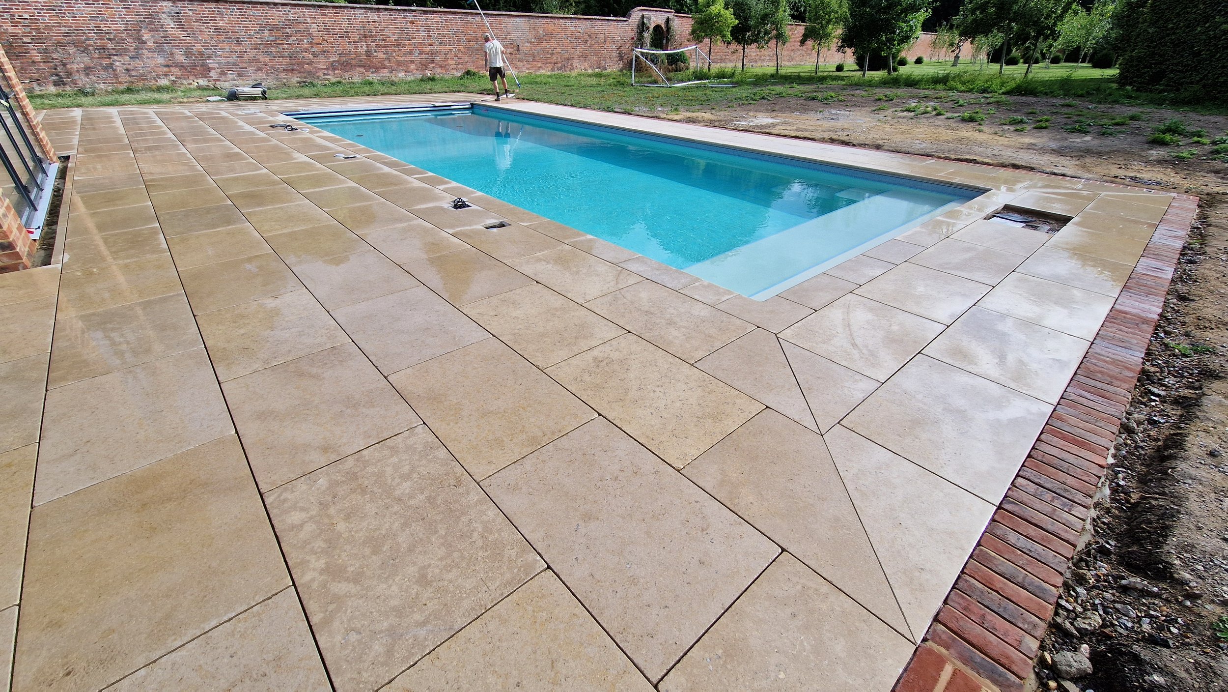 Backyard with a swimming pool surrounded by beige stone pavers, a brick wall in the background, and some trees and grass on the side.