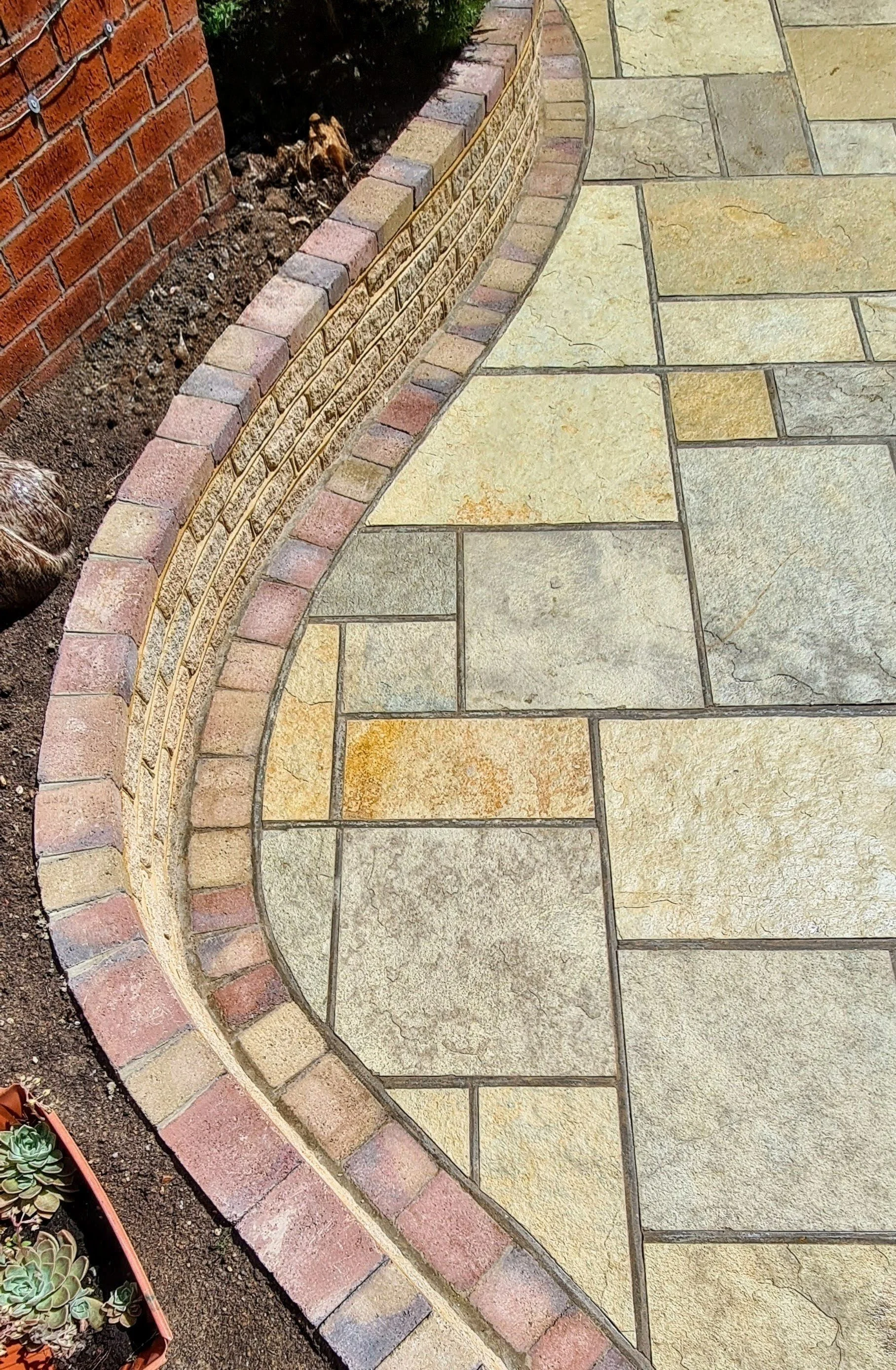 Curved brick border separating a garden bed with plants and soil from a paved walkway with large beige and gray stone tiles.