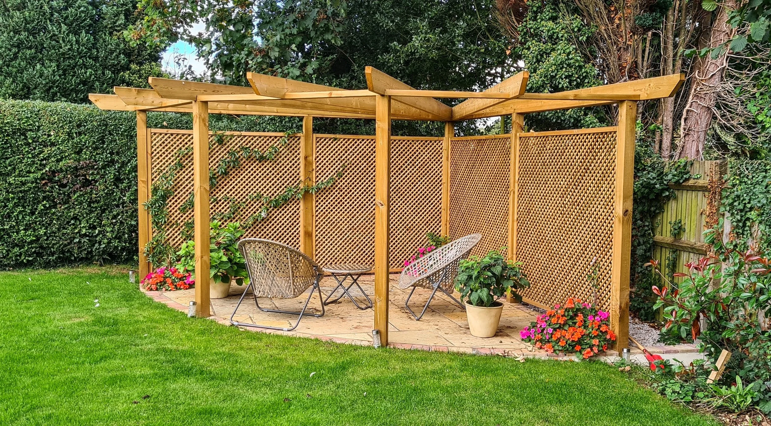 A wooden outdoor pergola with lattice side panels, situated on a brick patio in a garden. There are potted plants and chairs underneath, and greenery including bushes and trees surrounds the area.