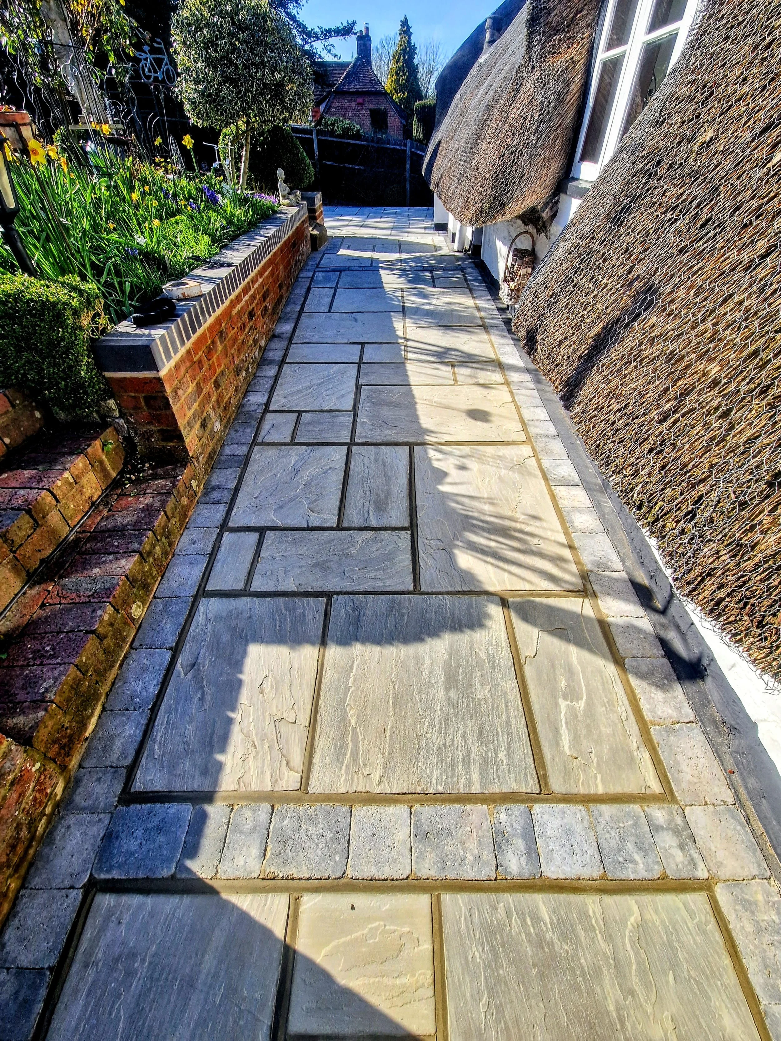 Stone patio pathway leading to a garden and brick house with thatched roof.