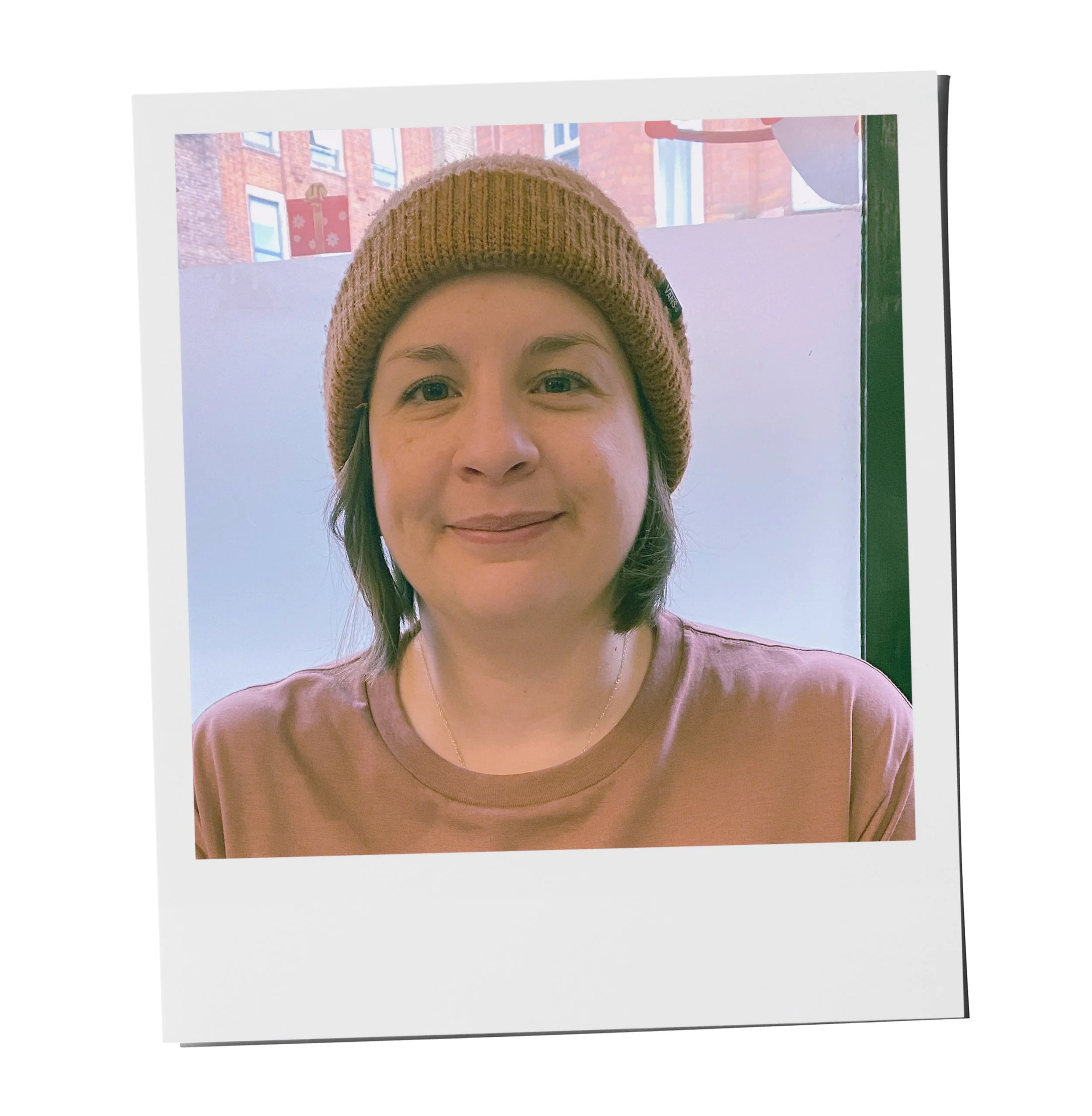 Partial view of a person wearing a brown knit beanie and a pinkish-brown shirt, smiling, with a background showing windows and a brick building outside.