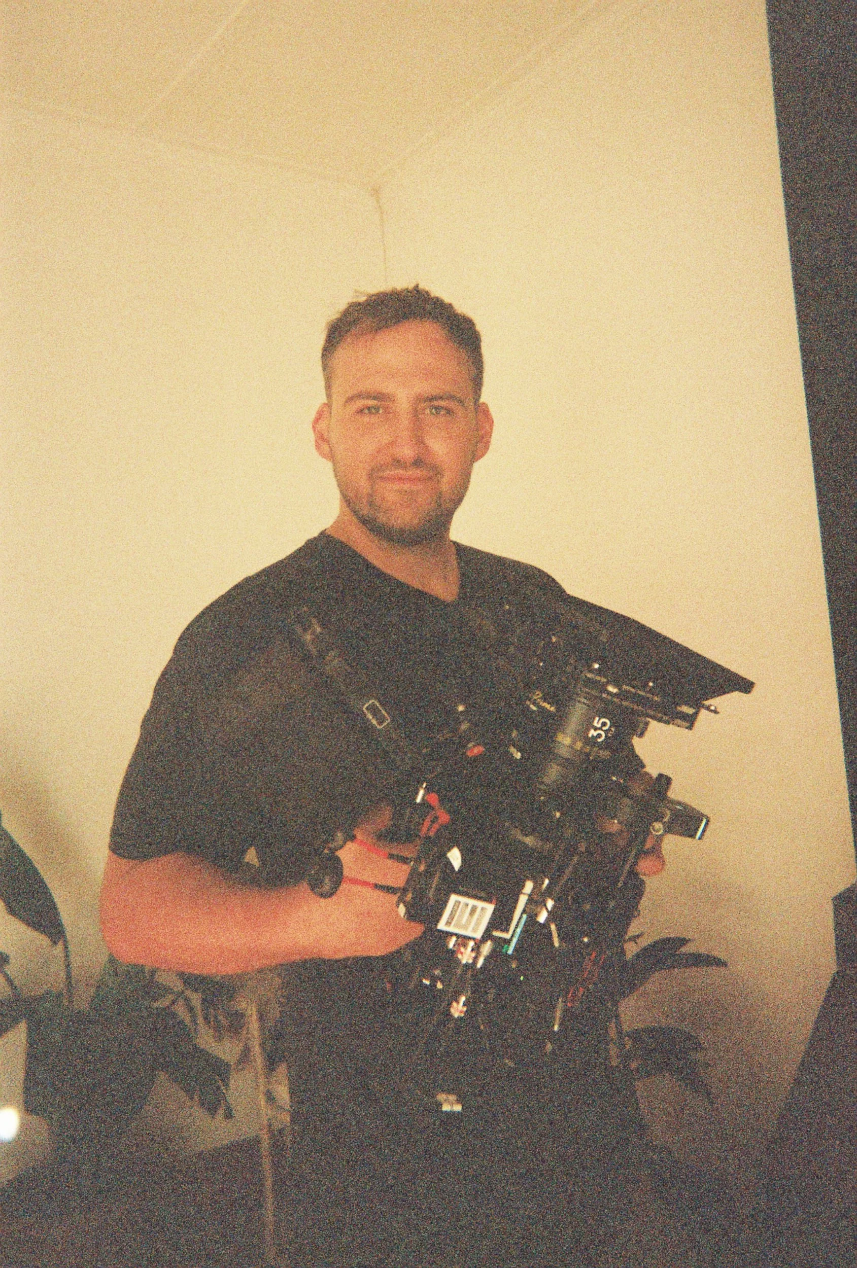 A man with short dark hair and stubble, wearing a black t-shirt, holding a professional film or photographic camera, standing in a room with beige walls and a plant in the background.