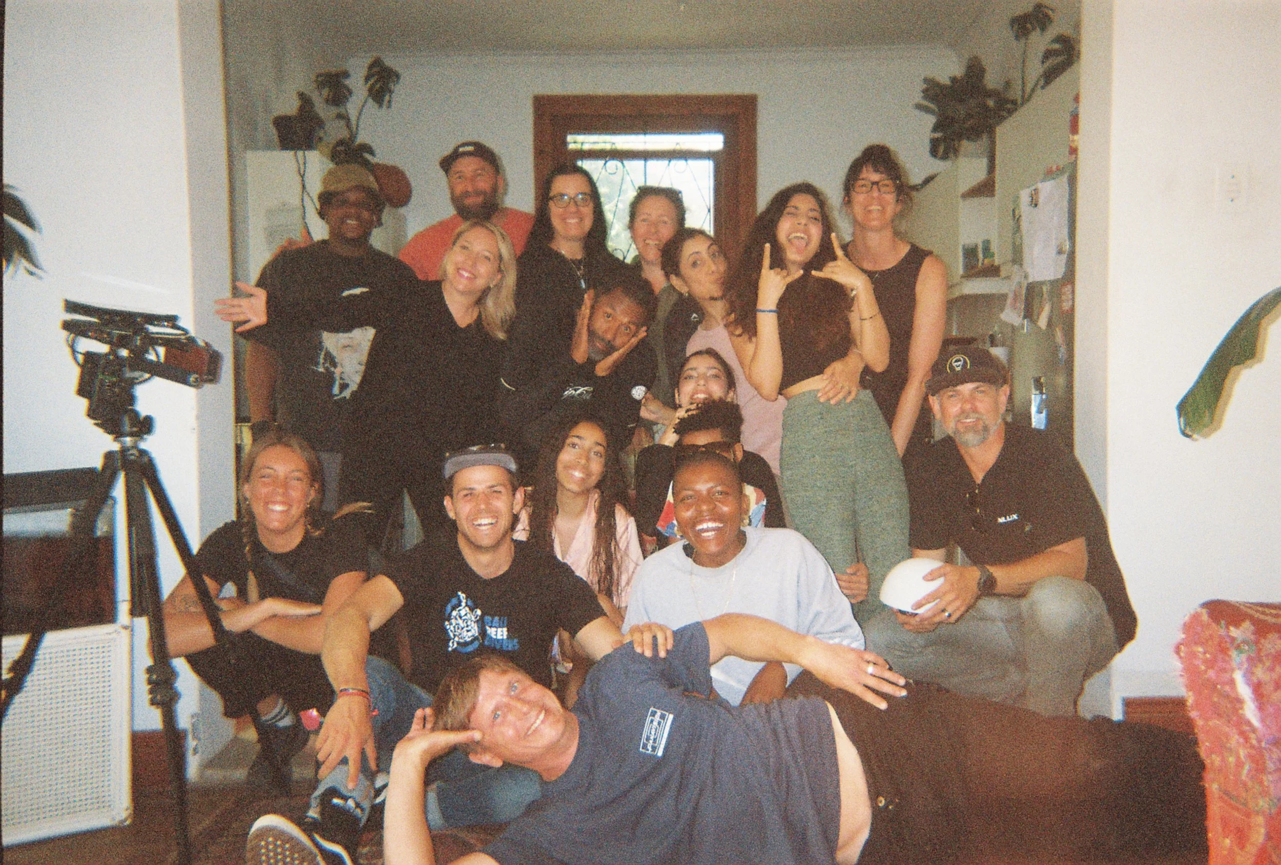 A group of diverse people posing together indoors, smiling and making playful gestures, with some standing and others sitting or lounging on the floor.