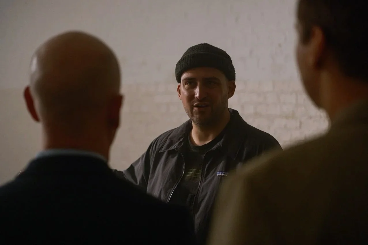 A man wearing a black beanie and jacket talking to two other people in a room with a plain wall.