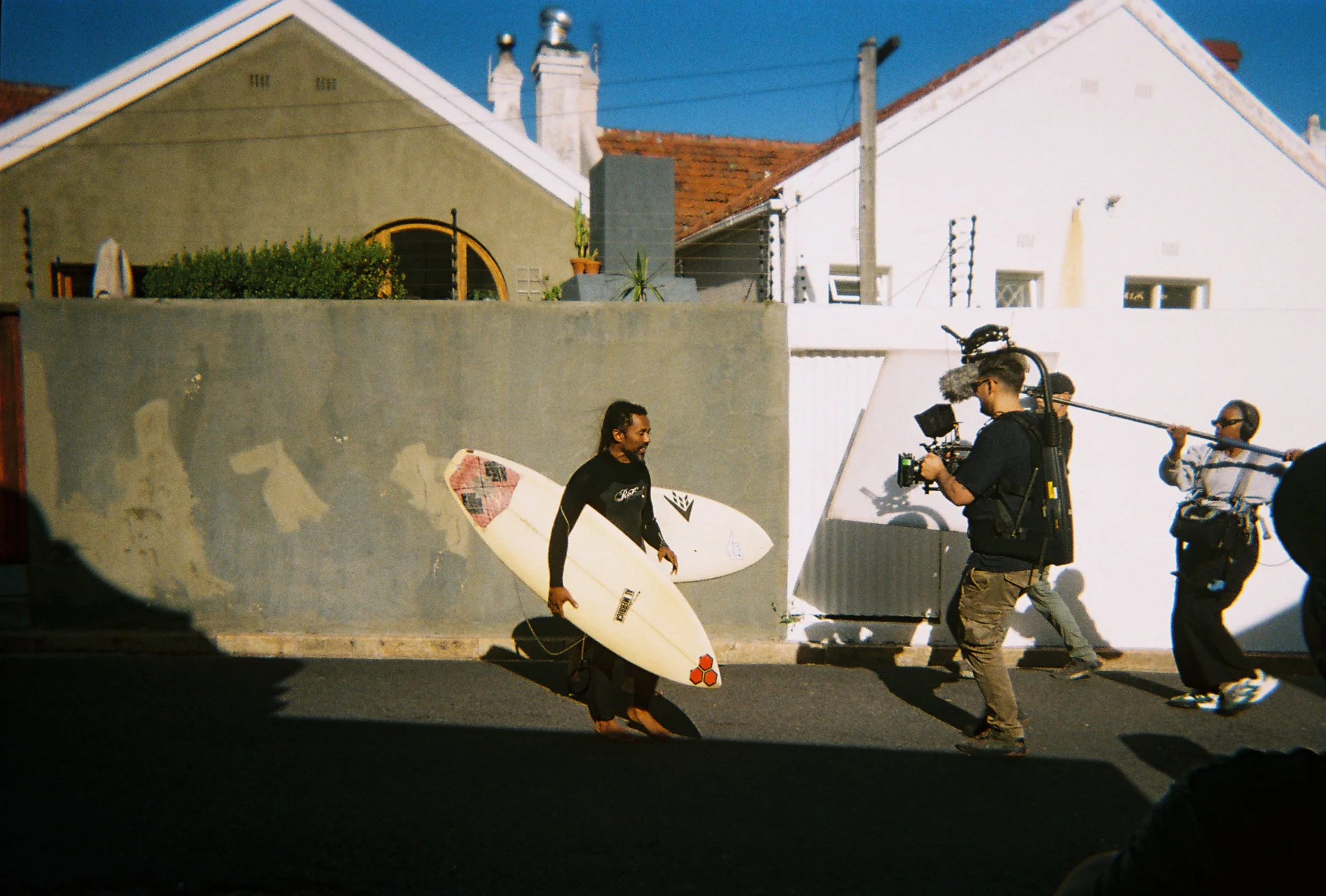 A person holding a surfboard stands next to a group of people filming or taking pictures. The scene appears to be outdoors on a sunny day in a neighborhood with houses and utility poles visible in the background.