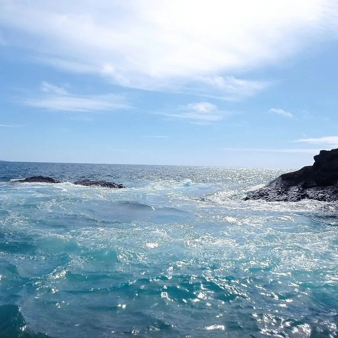 Ocean waves crashing against rocks under a blue sky with scattered clouds.