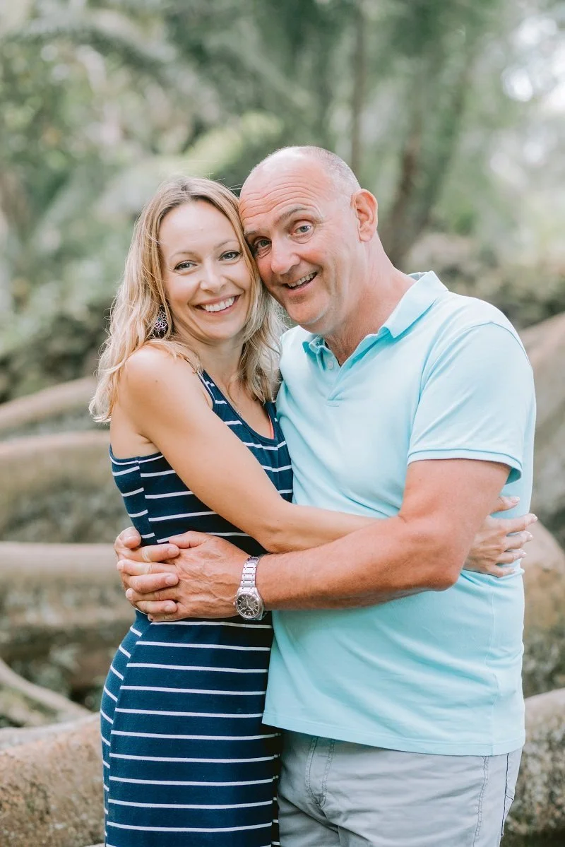 Melanie and Paul  hugging outdoors in a park, with trees and greenery in the background.