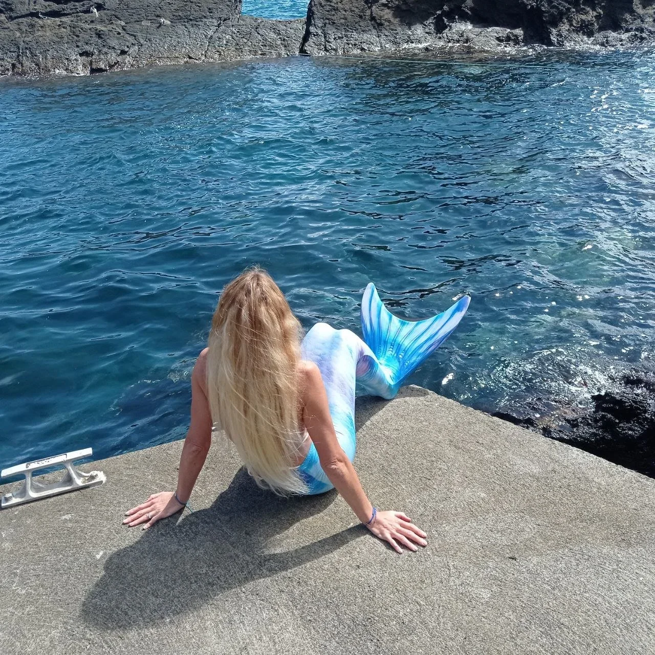 White Feathers Woman sits on a concrete dock by the water, facing away from the camera, with her hands on the ground supporting her. The water is calm and blue, and the background features a rocky shoreline.