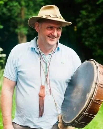 Paul/Grey Elk, smiling, holding a drum outdoors with greenery in the background.
