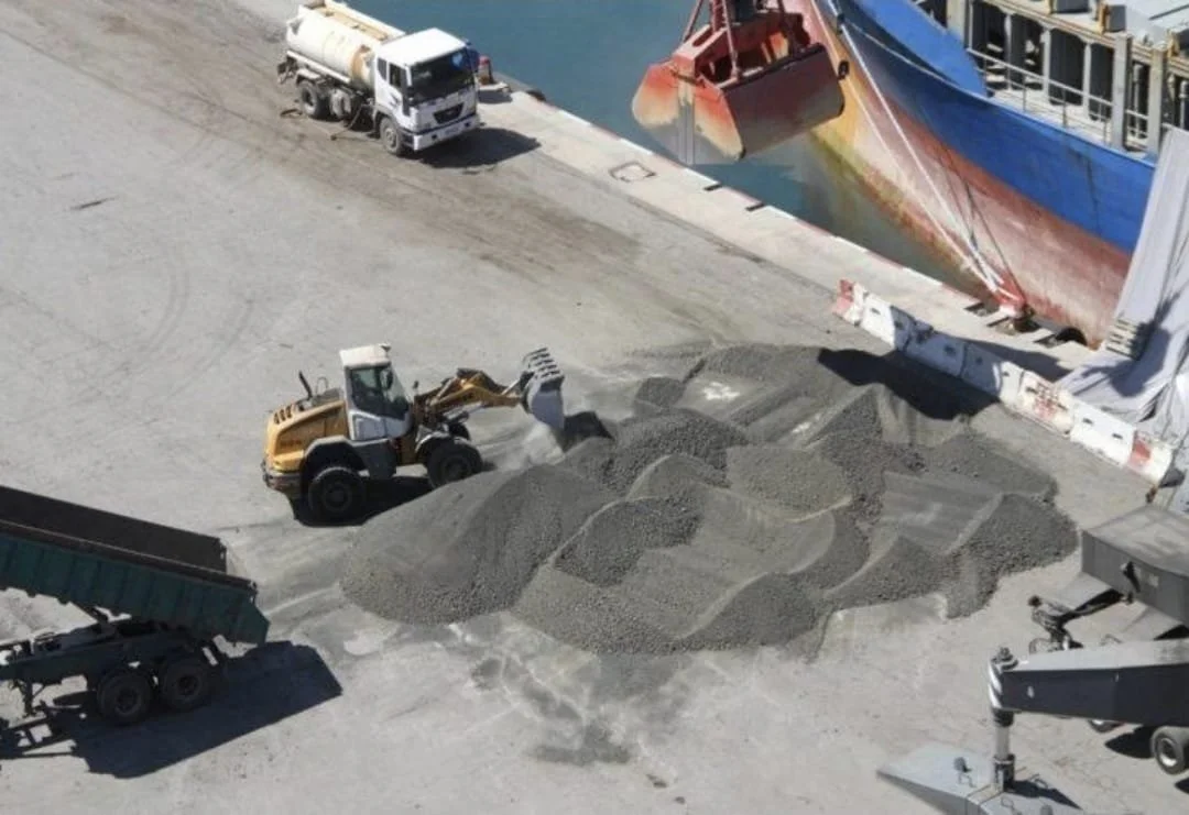 Vue aérienne d'un chantier naval avec une grue déchargeant du ciment , un bulldozer déplaçant des matériaux, un camion, et un bateau au quai.