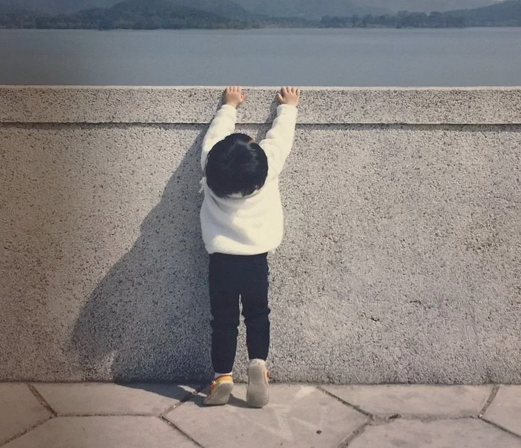 A young child with black hair, wearing a white sweater, black pants, and sneakers, standing on a paved surface and reaching up to hold onto the top of a concrete barrier while looking over it at a body of water.