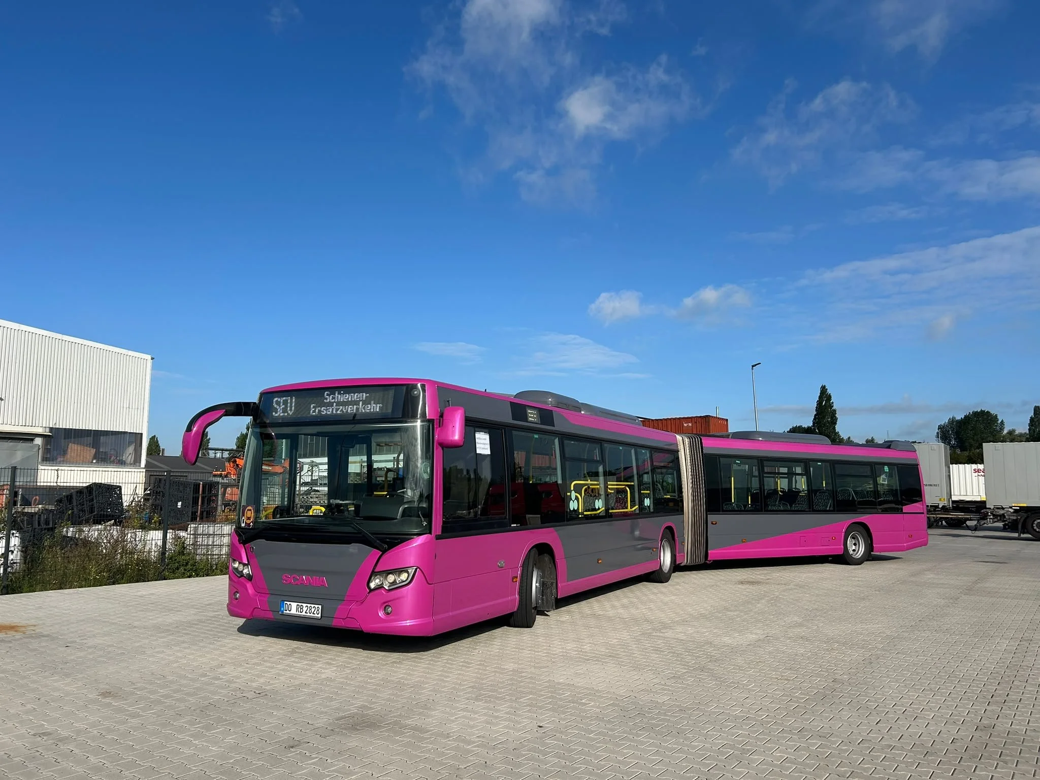Ein pinker Gelenkbus steht auf einem gepflasterten Parkplatz vor einem Industriegebiet. Der Himmel ist größtenteils klar mit wenigen Wolken.