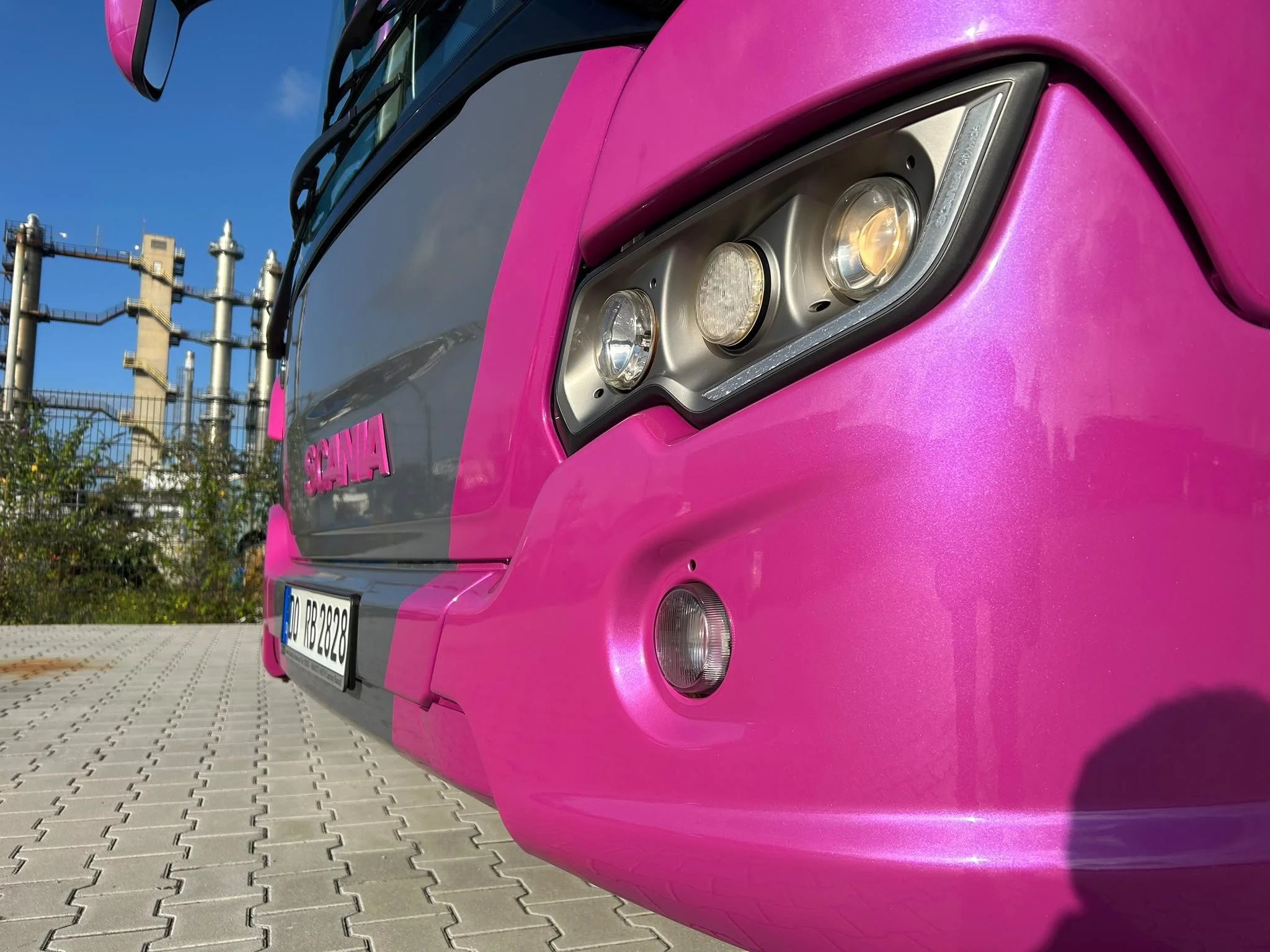 Naheaufnahme der Frontseite eines pinkfarbenen Autos, das auf einer gepflasterten Fläche steht, mit einer Industrieanlage im Hintergrund und einem blauen Himmel.