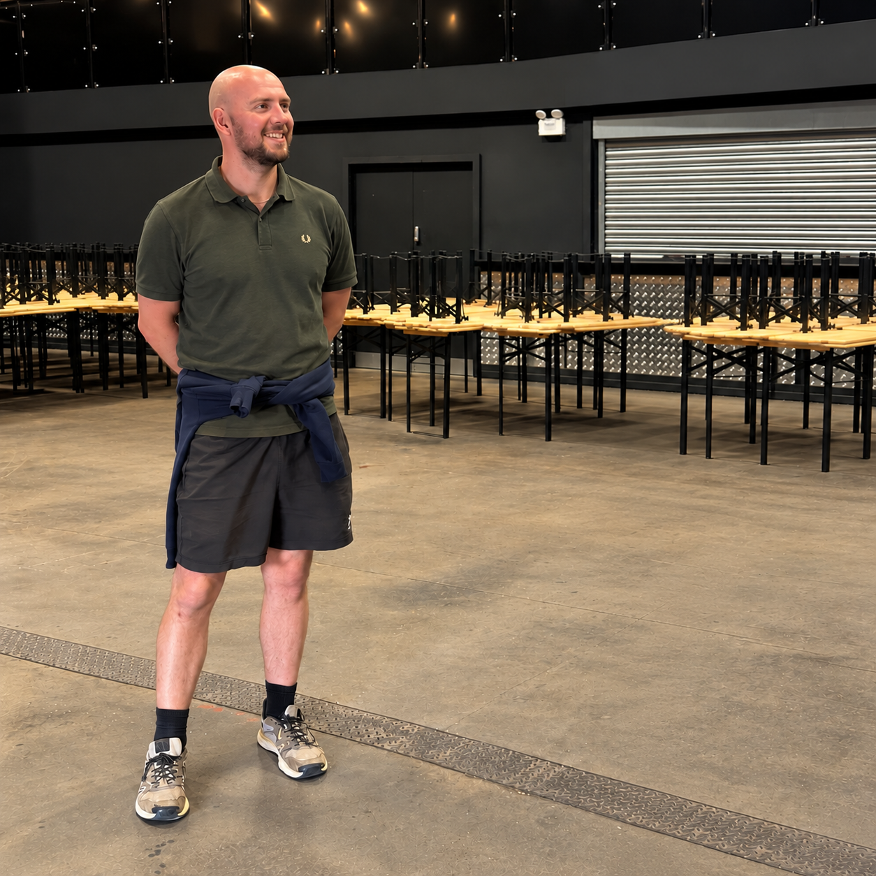 A smiling man with a beard, wearing a green polo shirt, black shorts, and sneakers, standing in an empty room with folded tables and chairs stacked along the wall.