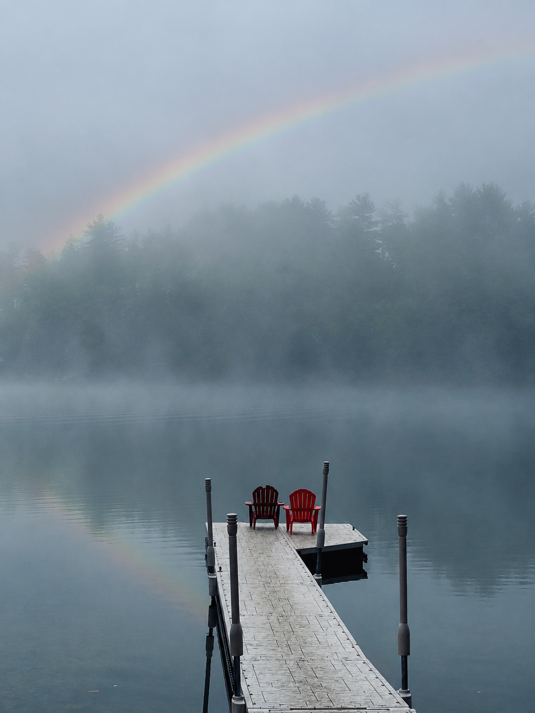 A foggy lakeside scene with a wooden dock extending into the water, two red Adirondack chairs on the dock, and a quiet lake with a distant tree line. A rainbow arcs across the sky.