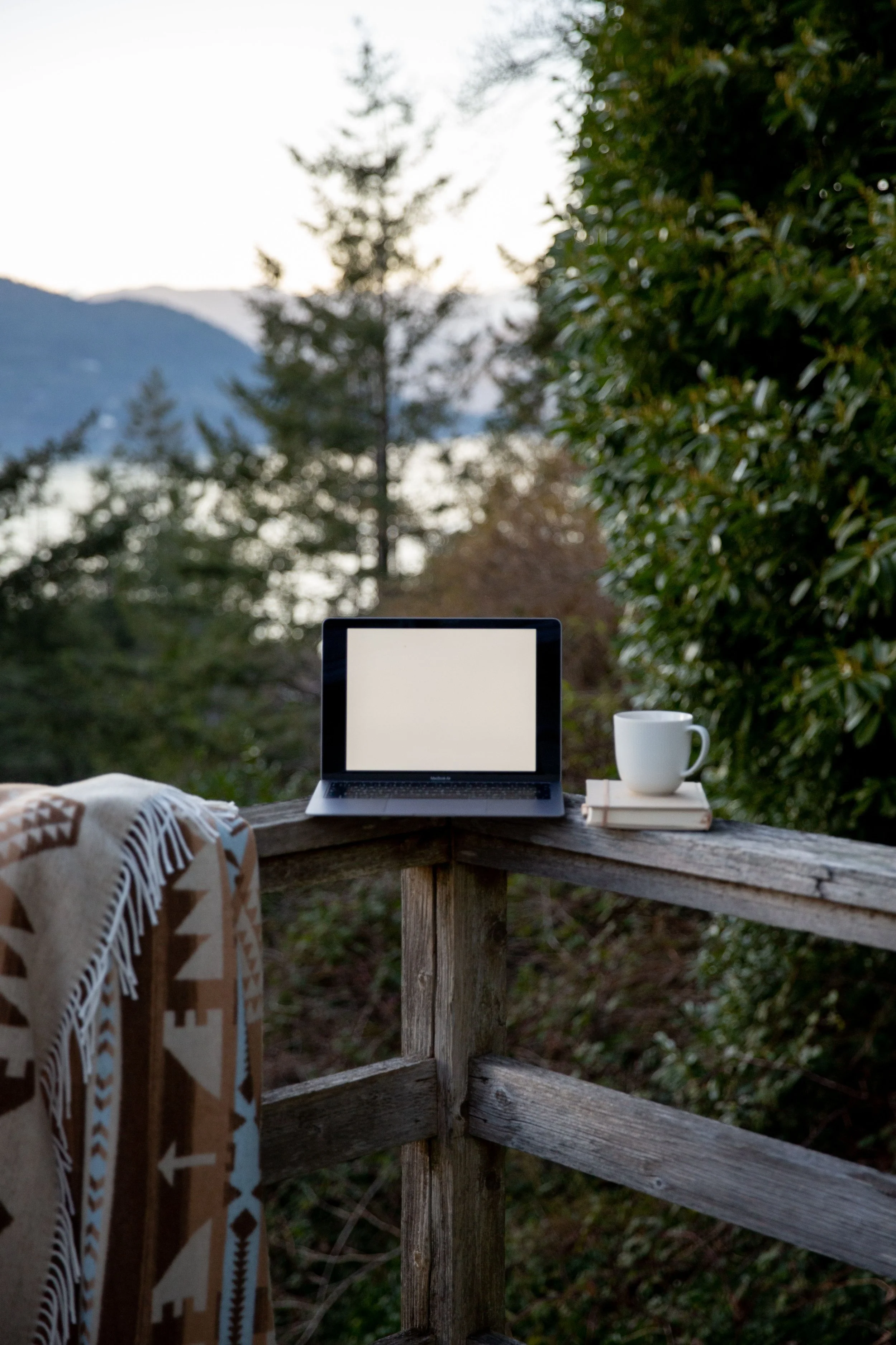 A laptop on a wooden railing outdoors with a white coffee mug and a folded blanket nearby, and trees with mountains in the background.