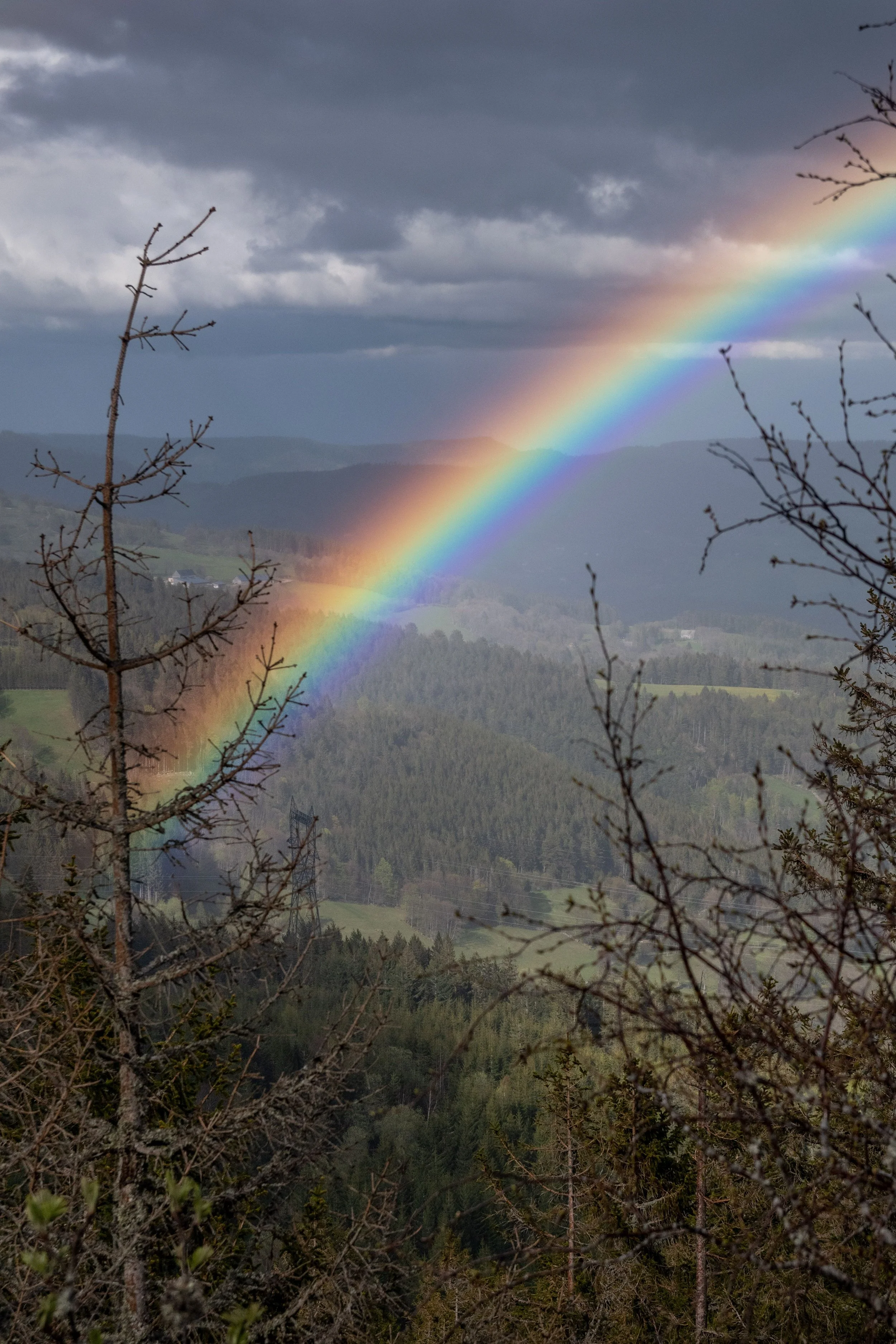 A rainbow arching across a cloudy sky over a hilly landscape with trees and some houses in the distance.