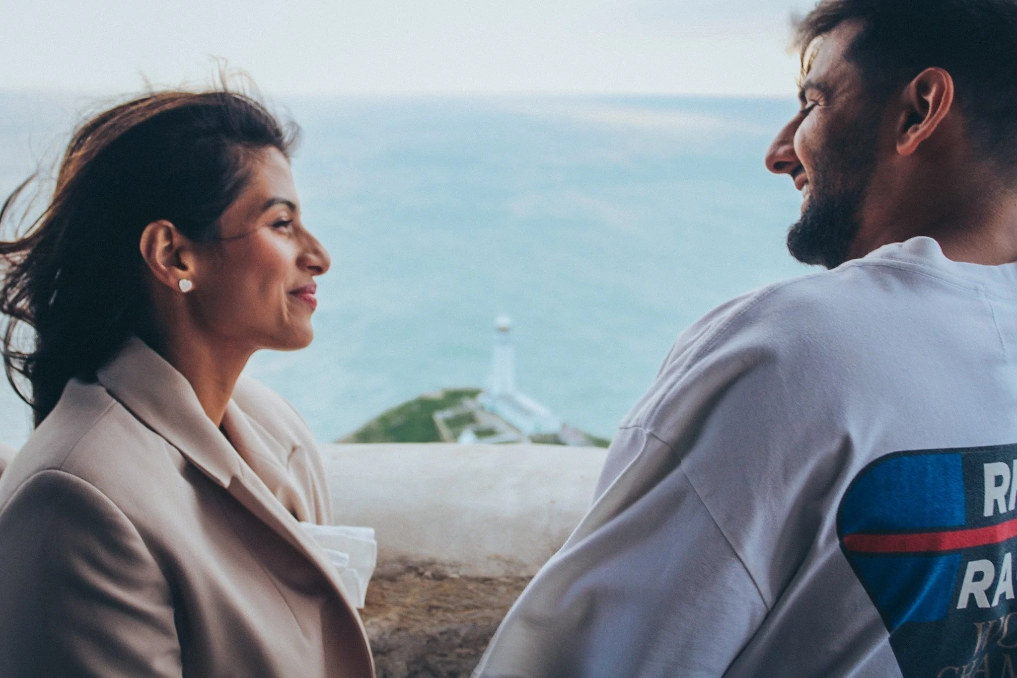 A couple smiling at each other on a clifftop by the sea, moments after a proposal