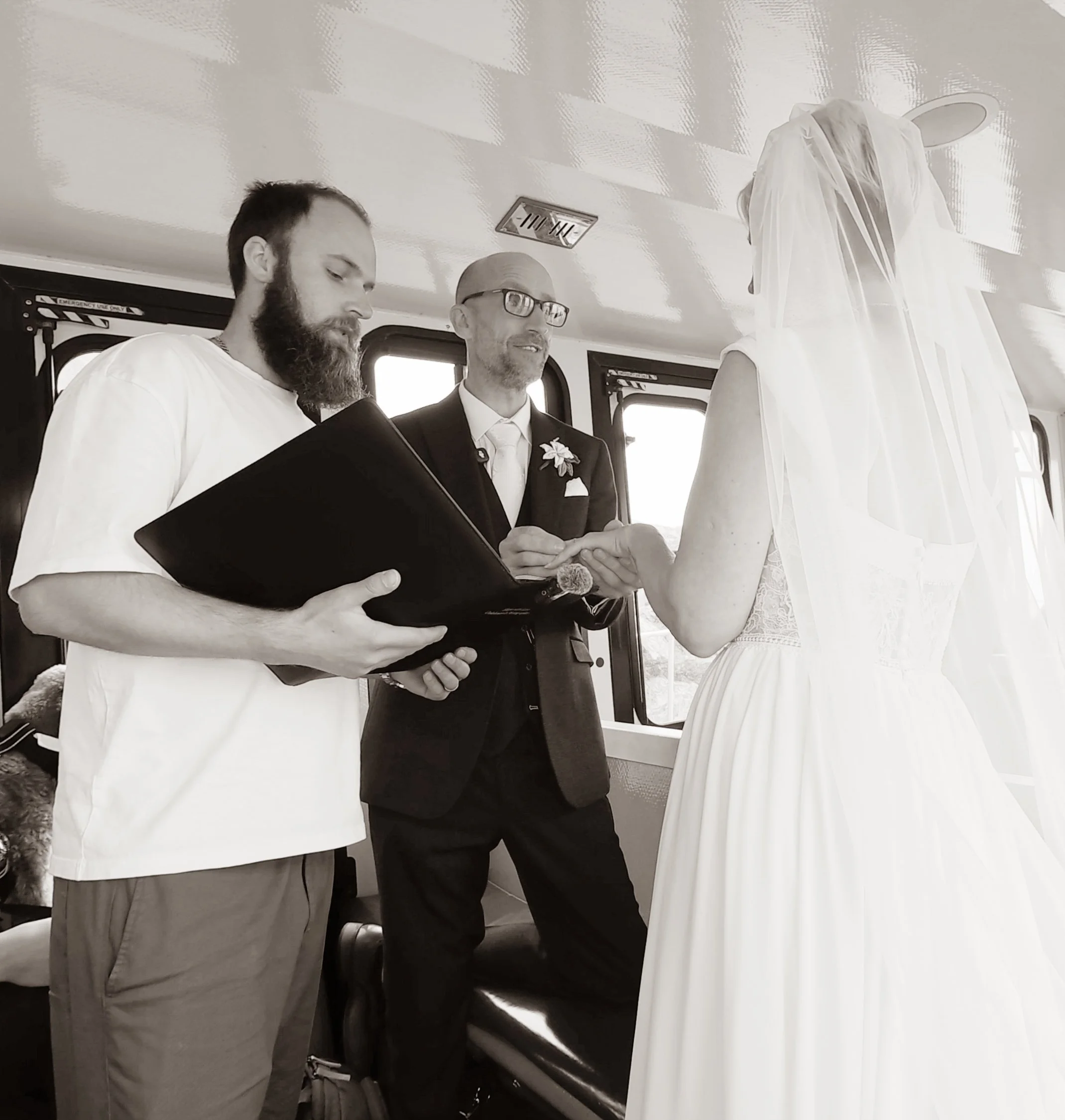 Leon officiating as a couple exchange rings inside a carriage, in black and white