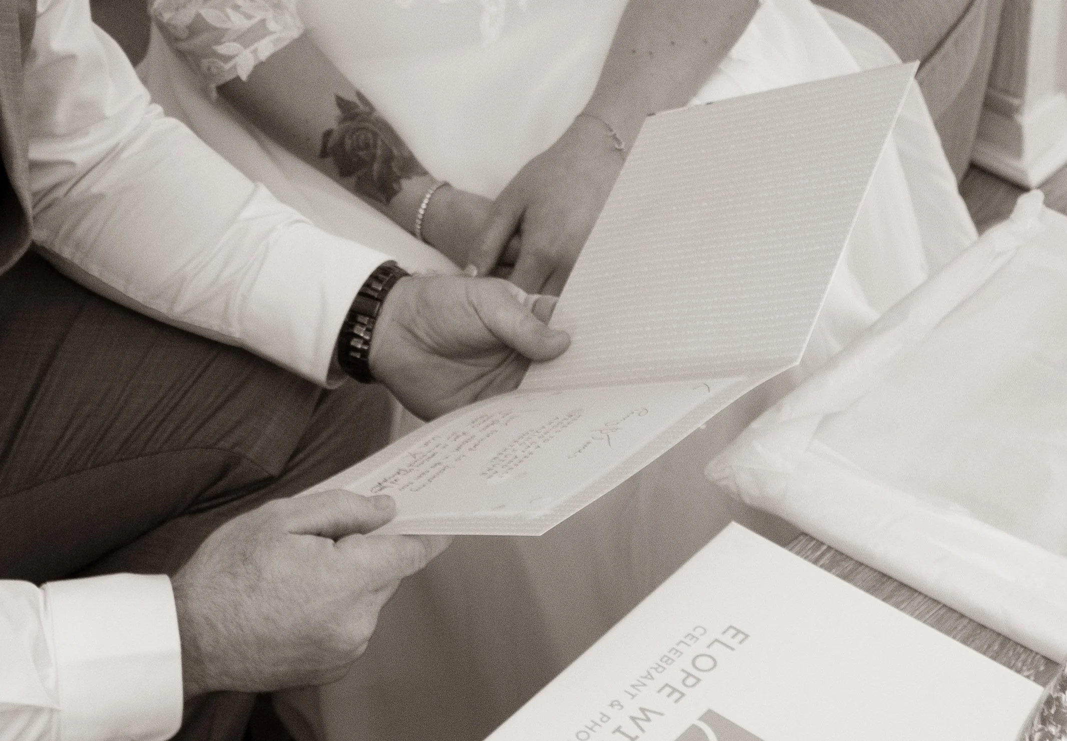 A couple's hands holding a card in front of a gift box, in black and white