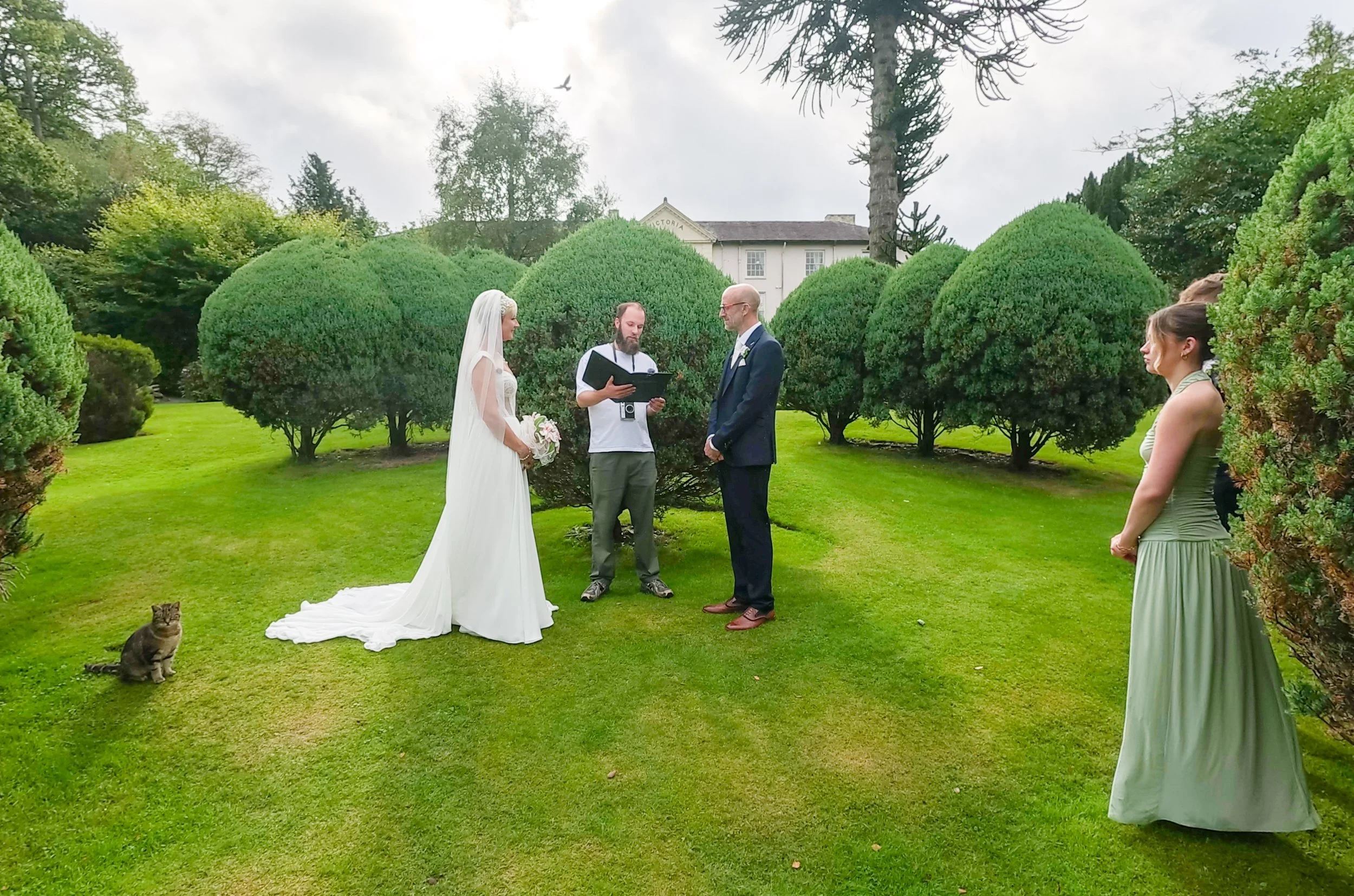 A garden ceremony on a lawn among clipped topiary, with a bridesmaid watching